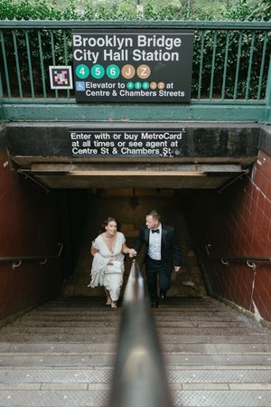 A couple in formal wedding attire descends the stairs at Brooklyn Bridge City Hall subway station entrance.