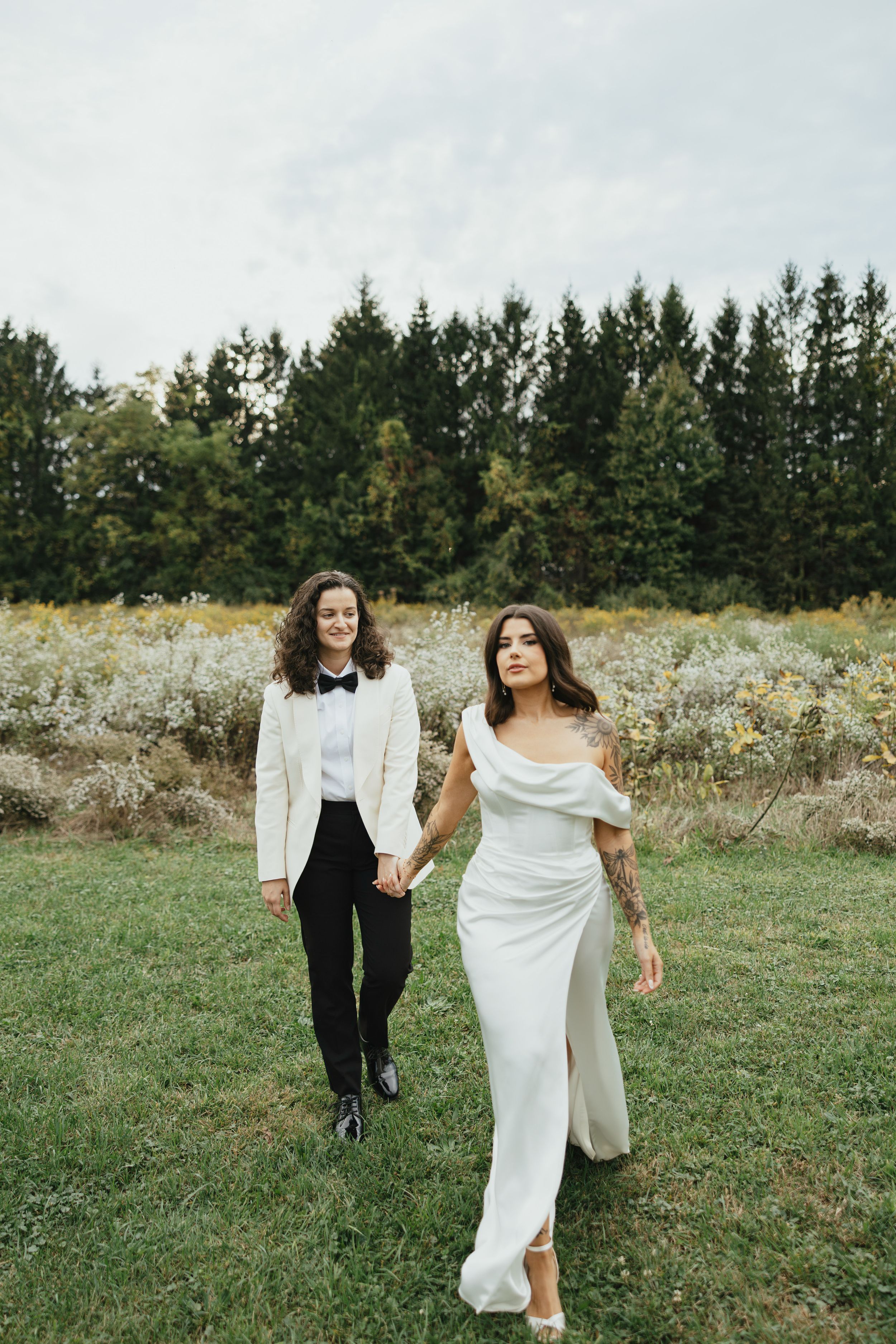 A couple in wedding attire walks together through a wildflower meadow with evergreen trees in the background.