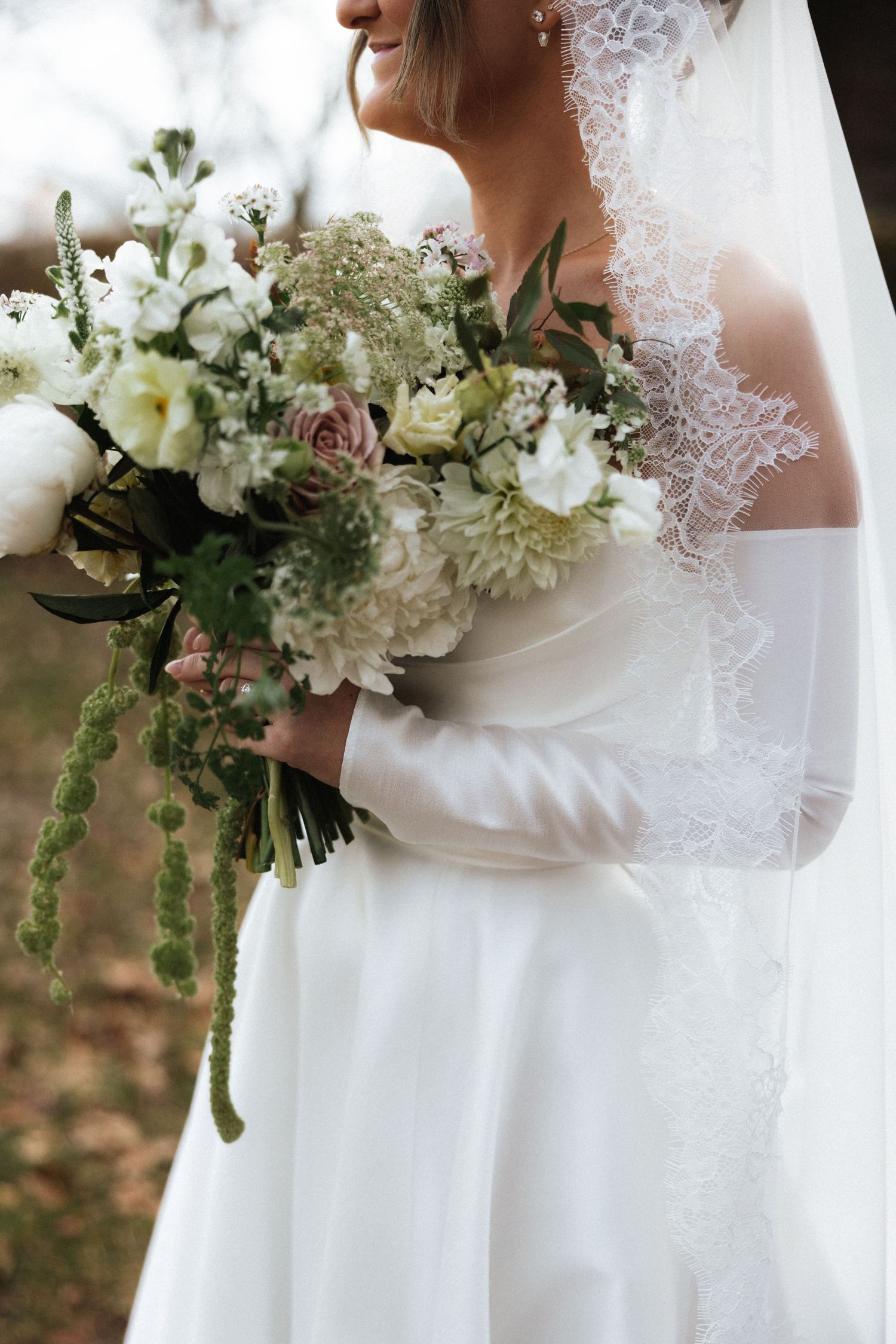 Bride in an off-shoulder white gown and lace veil holds a lush bouquet of white and blush flowers.