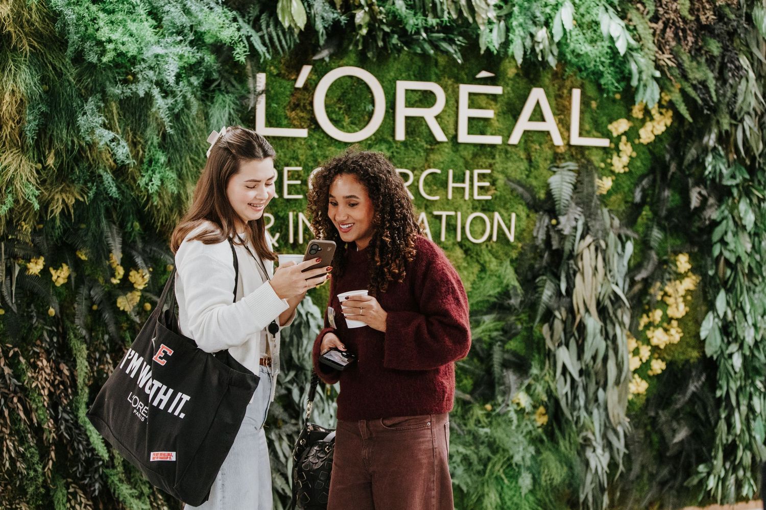A person in a burgundy sweater stands in front of a lush green plant wall with L'Oreal branding.