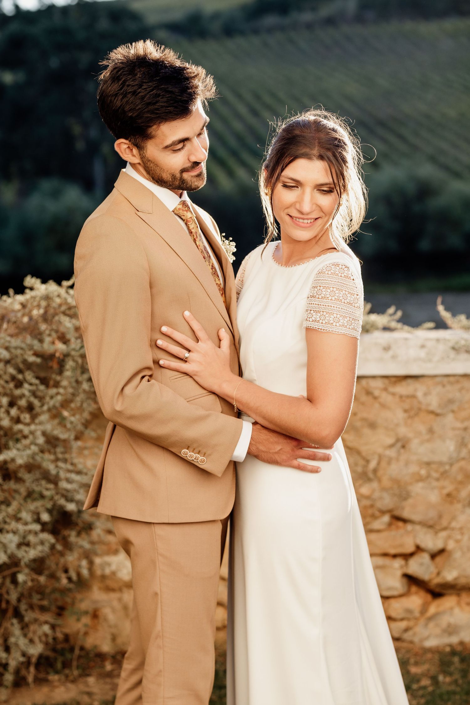 Elegant wedding couple in tan suit and lace dress pose together against stone wall.