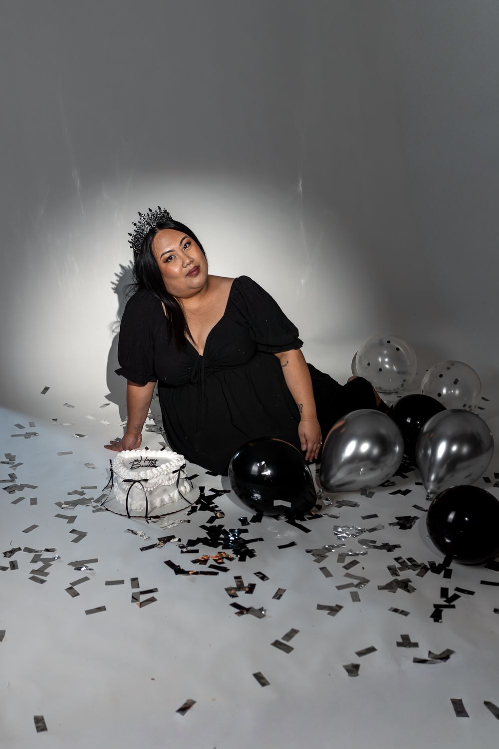 A person in black attire lies on a white floor surrounded by scattered black confetti and balloons in a moody setting.