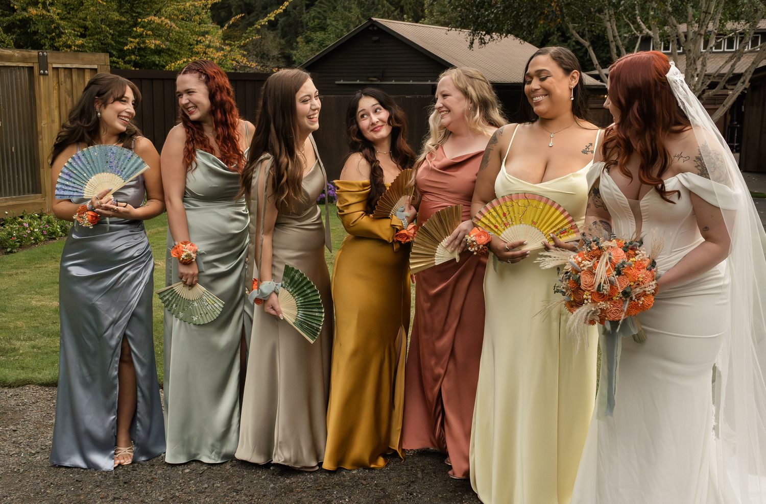 Bridesmaids in soft pastel and earth-toned dresses stand together holding colorful autumn bouquets at an outdoor wedding.