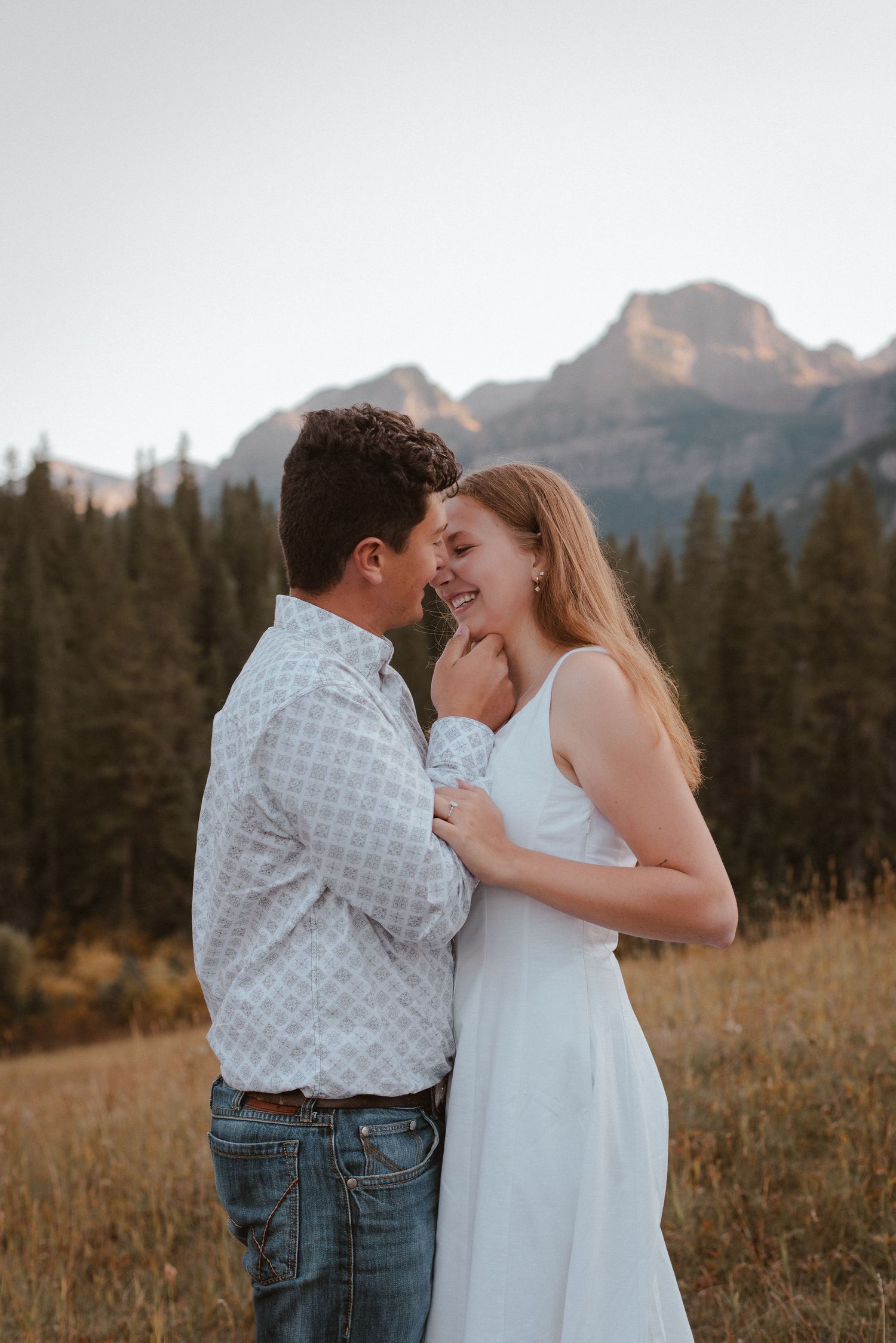 A couple shares a romantic moment in a mountain meadow with evergreen trees in the background.