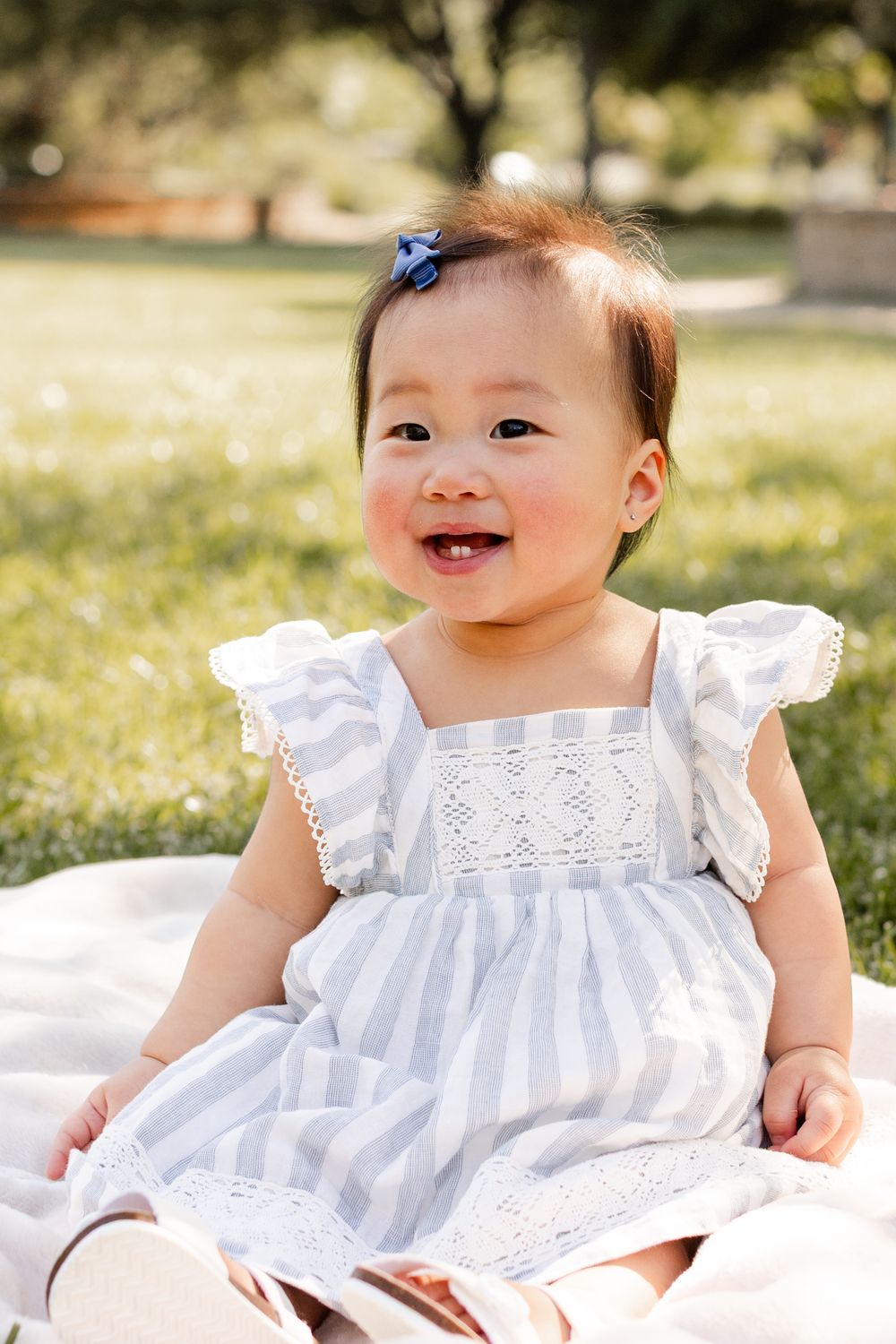 A baby wearing a white lace dress sits and smiles on grass during an outdoor photo session.
