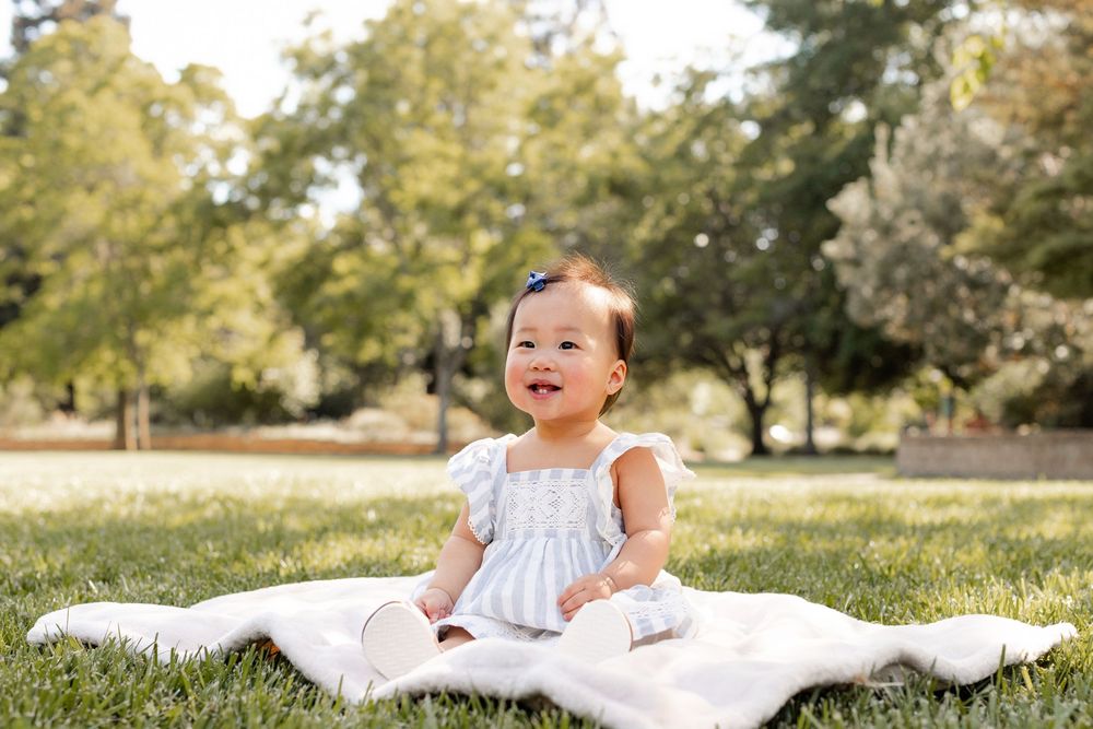 A baby in a white dress lies on a blanket in a grassy park setting during a sunny day.