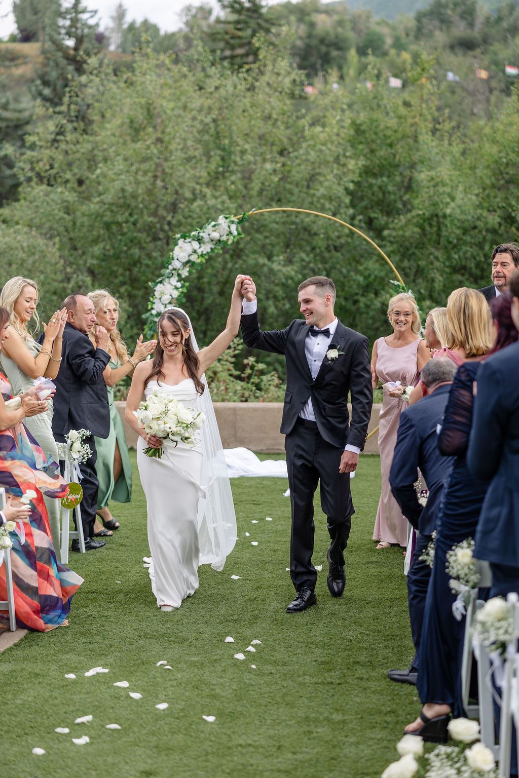 A wedding ceremony on a green lawn with guests watching the couple walk down the aisle under floral decor.