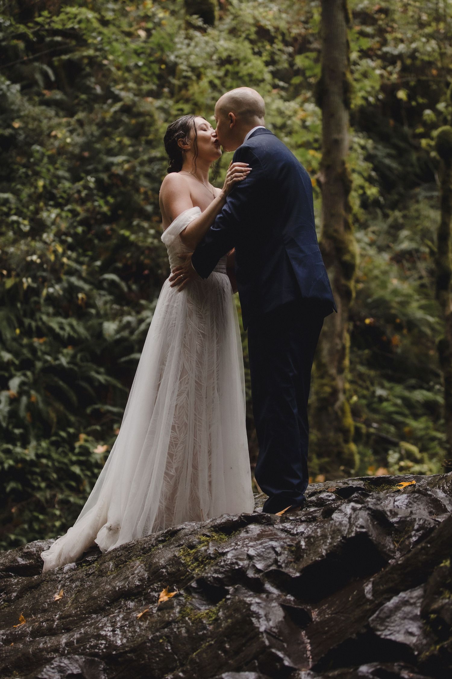 A couple in wedding attire share an intimate moment while standing on rocks in a wooded forest setting.