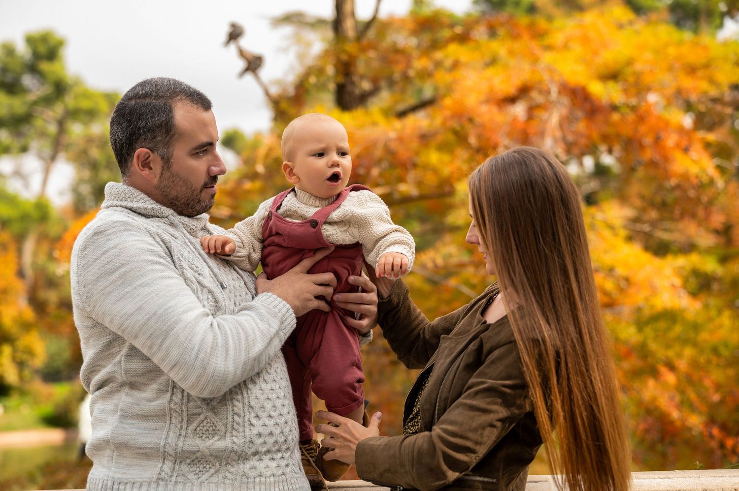 People in sweaters playfully tug at a football in a park with vibrant autumn foliage in the background.