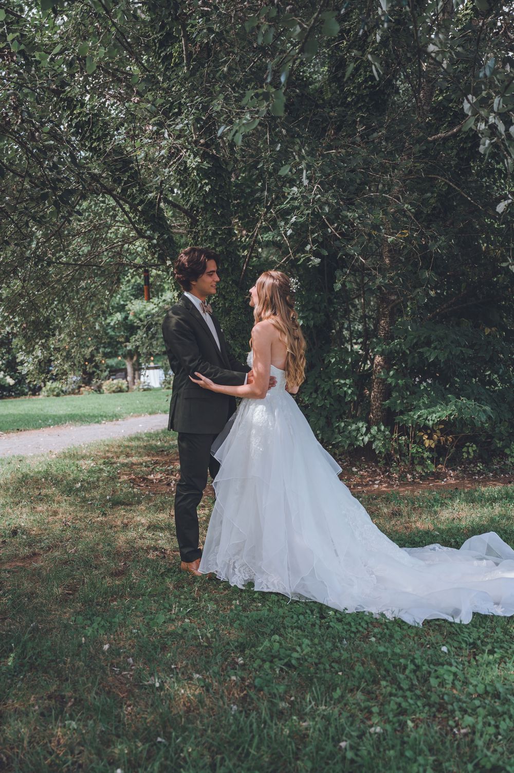 A couple in wedding attire stands together on grass beneath trees, with the bride wearing a flowing white gown with long train.