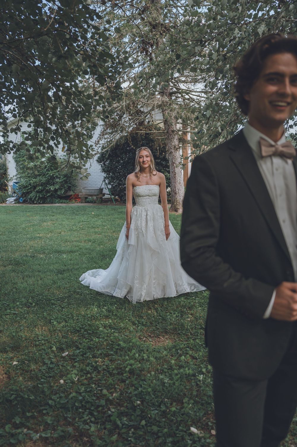 A bride in a white lace gown stands on grass while a groom in a black tuxedo is positioned in the foreground.