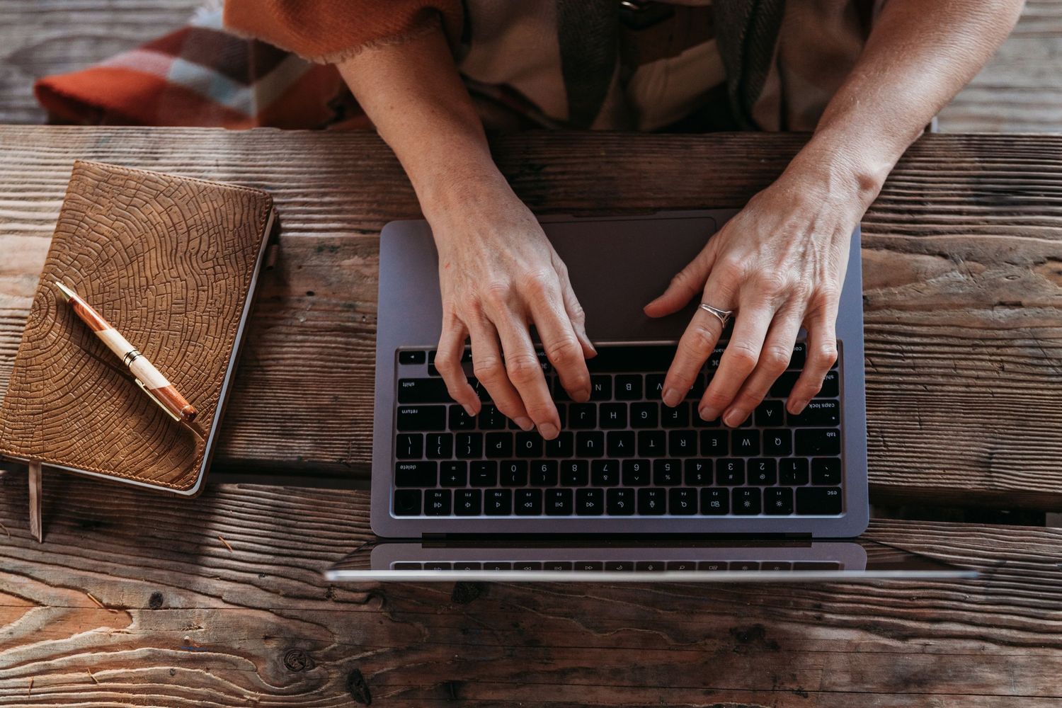 Hands typing on laptop keyboard on rustic wooden desk with brown leather notebook and pencil.