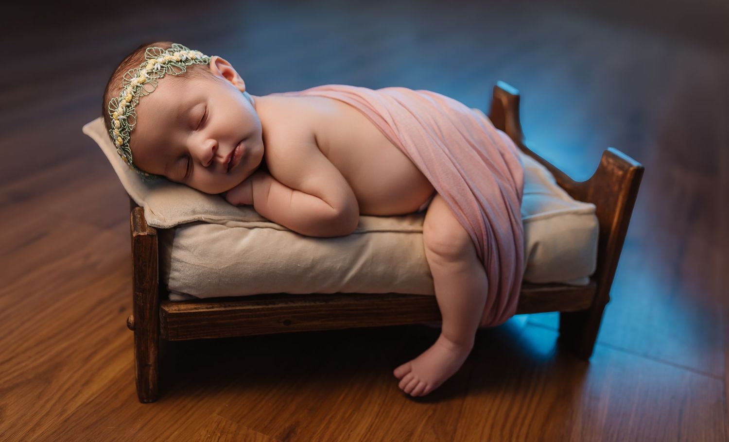 A sleeping newborn draped in soft pink fabric rests peacefully on a tiny wooden bed with a cushion.