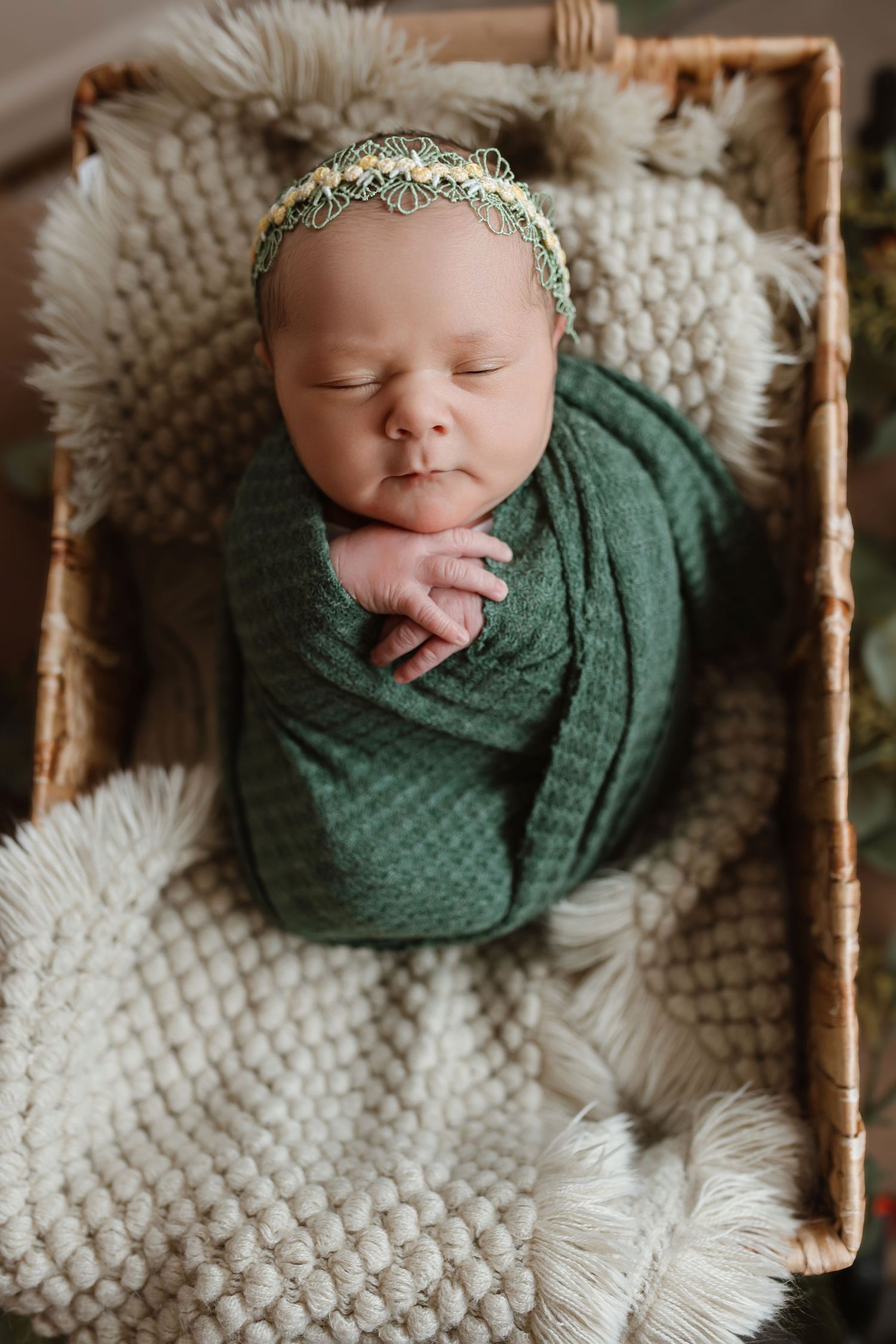 Sleeping newborn wrapped in emerald green swaddle on white textured blanket with delicate headpiece and rustic backdrop.
