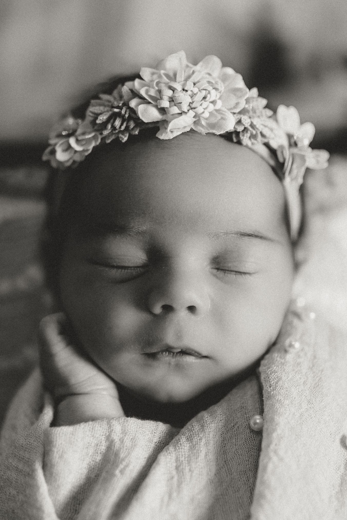 A black and white artistic photograph shows a sleeping baby wearing a delicate floral crown against a soft white blanket.