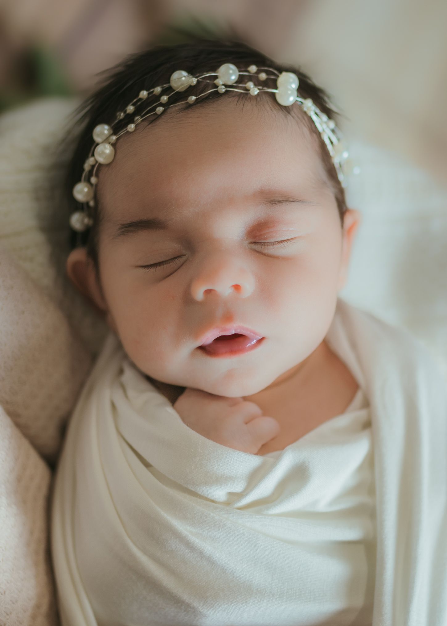 A sleeping newborn baby wearing a delicate white floral headband and wrapped in soft white fabric.