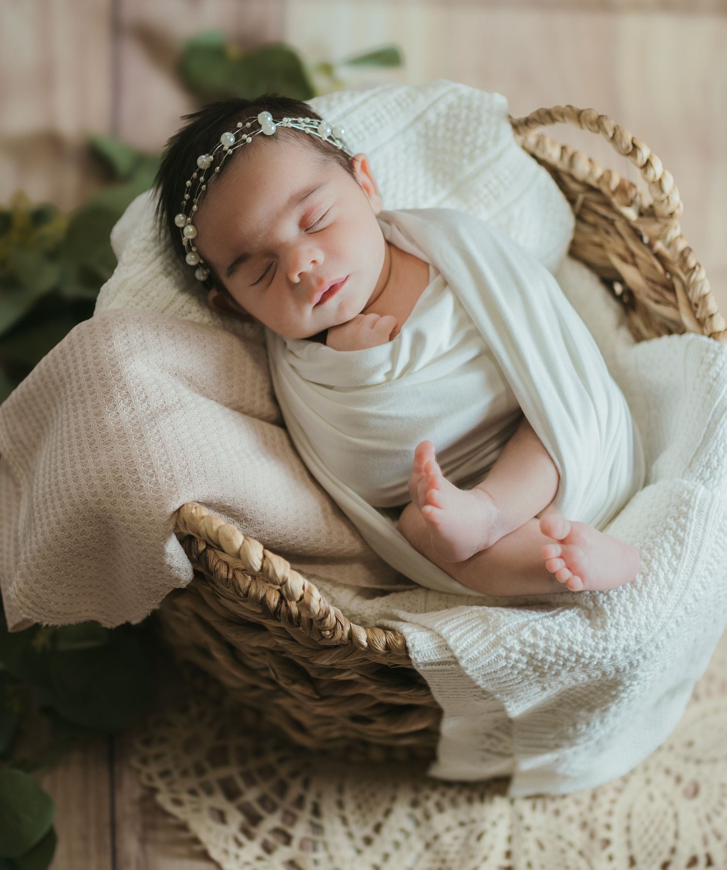 A sleeping newborn baby wearing white swaddle clothing lies peacefully in a woven basket on textured cream bedding.