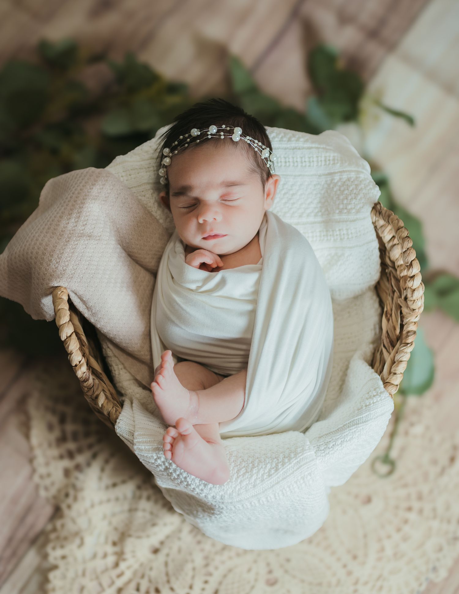 Newborn baby sleeps peacefully in white swaddle blanket on woven basket with lace backdrop in professional photo series.