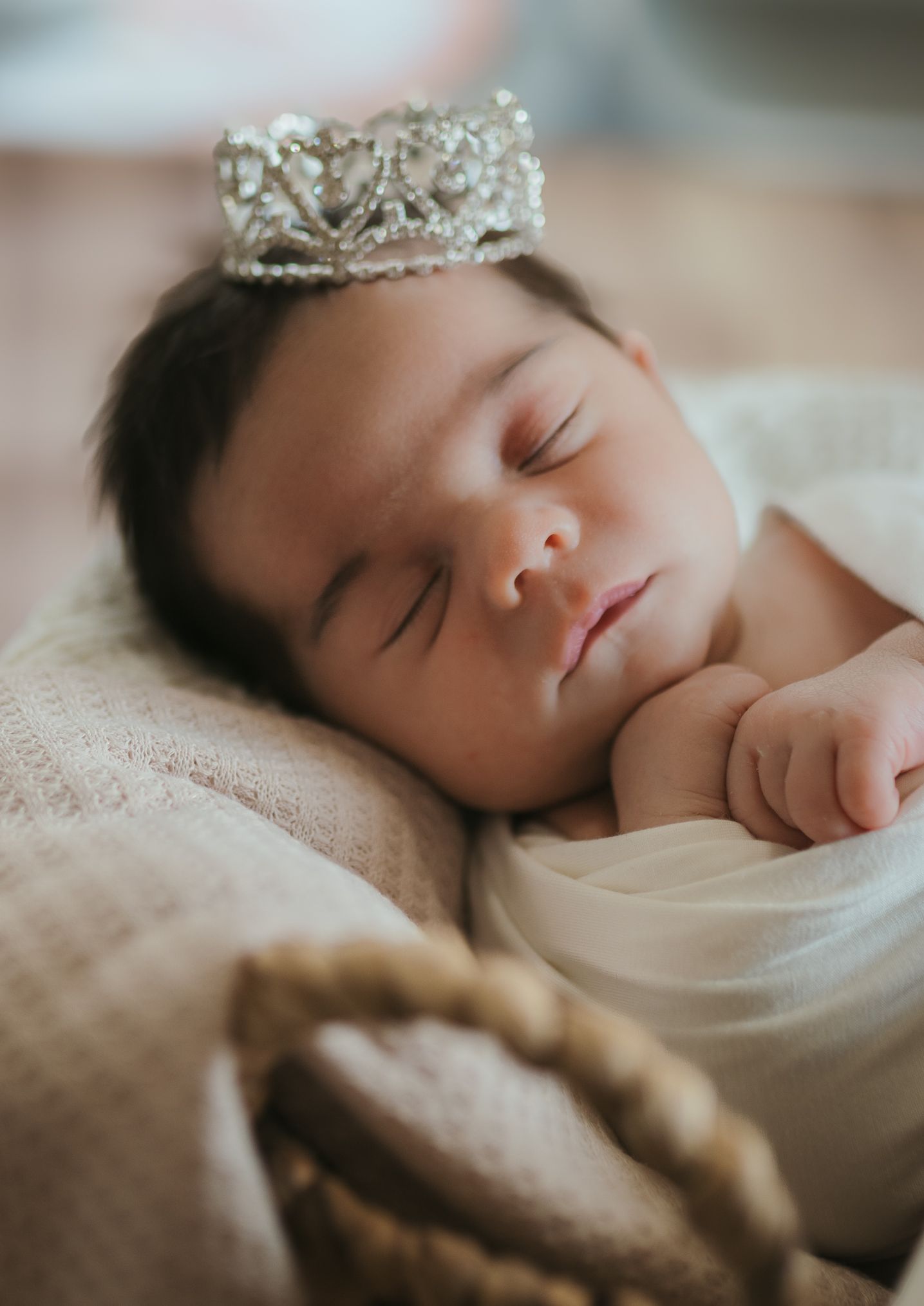 A sleeping baby wearing a delicate silver crown tiara rests peacefully on soft fabric.