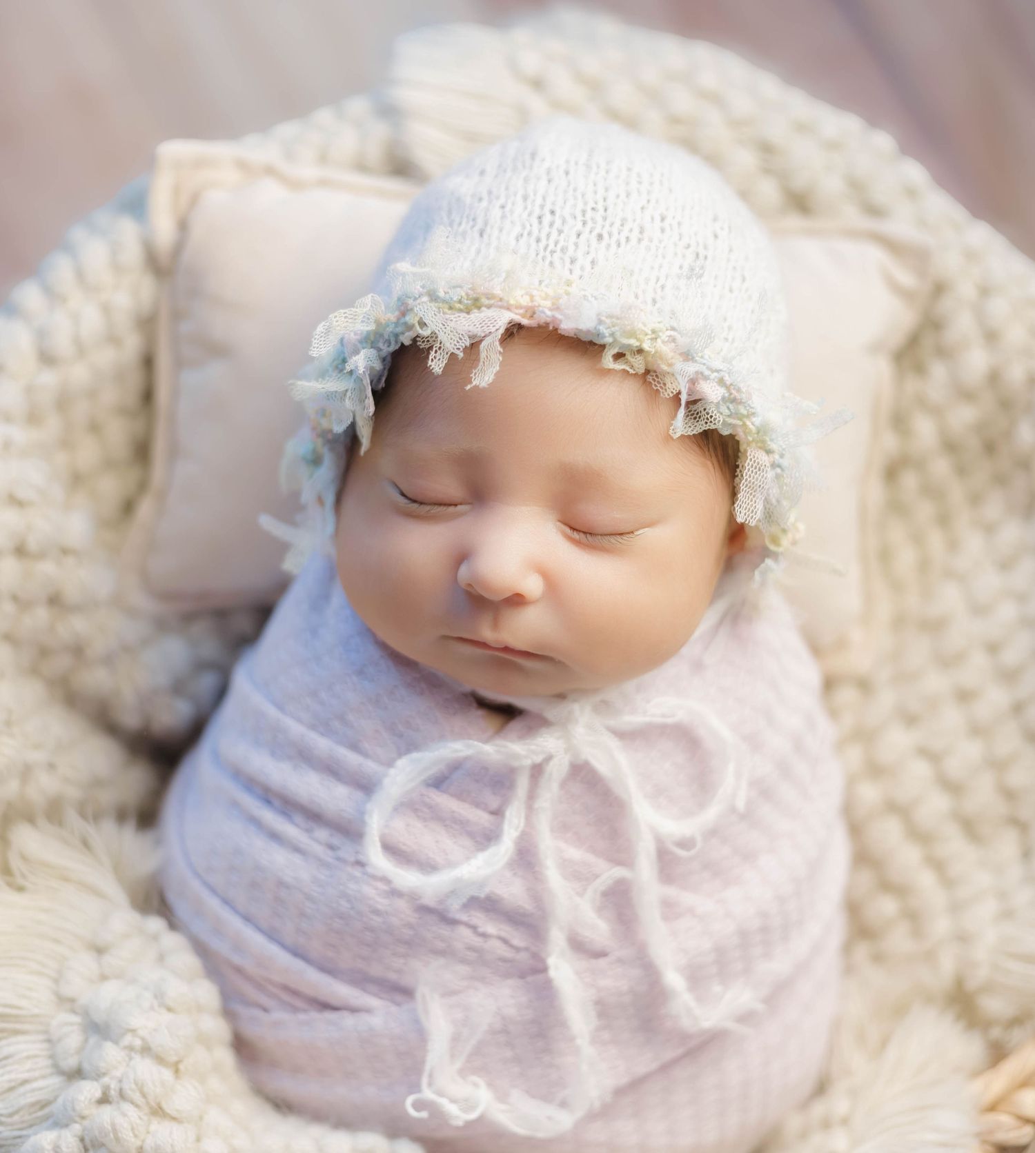 A sleeping newborn baby wrapped in soft pink fabric and delicate lace bonnet rests peacefully on a textured blanket.