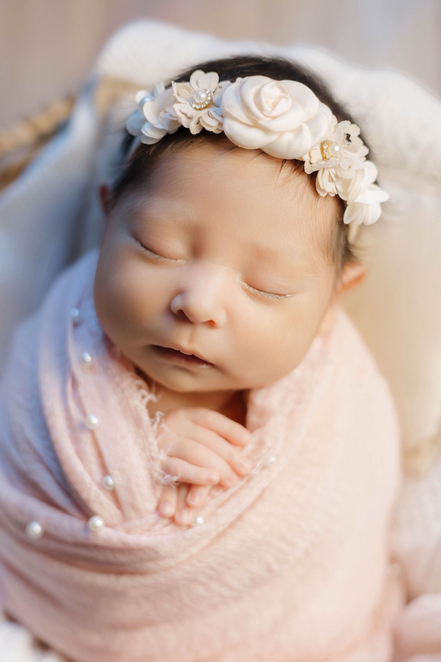 A sleeping newborn baby wrapped in soft pink fabric wearing a delicate white floral headband in a peaceful pose.