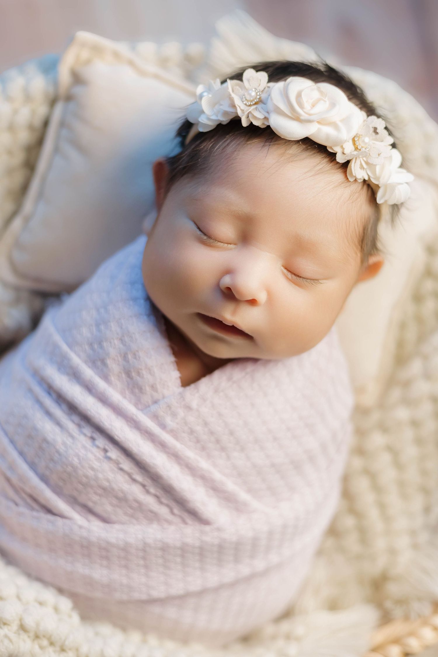 A sleeping newborn baby swaddled in soft white fabric wearing a delicate floral headband.
