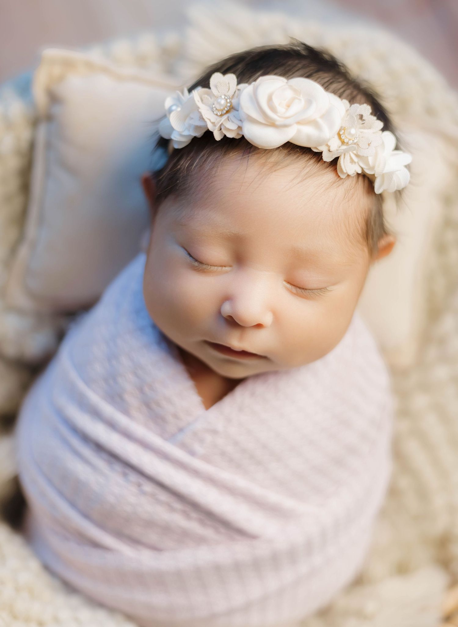 Sleeping newborn baby wrapped in soft pink swaddle wearing delicate white floral headband on textured cream blanket.