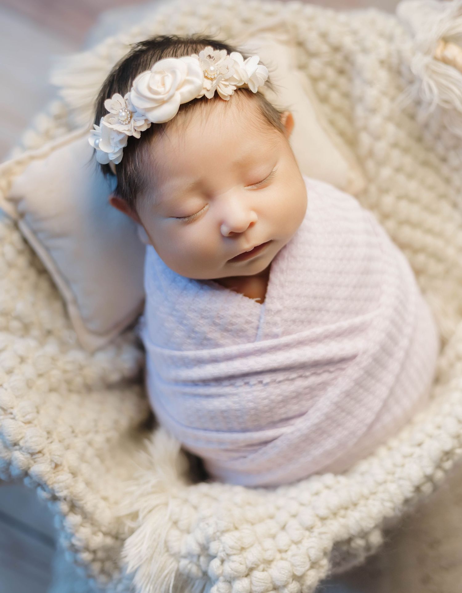 Peaceful newborn baby swaddled in pink fabric with white rose headband resting on cream textured blanket.