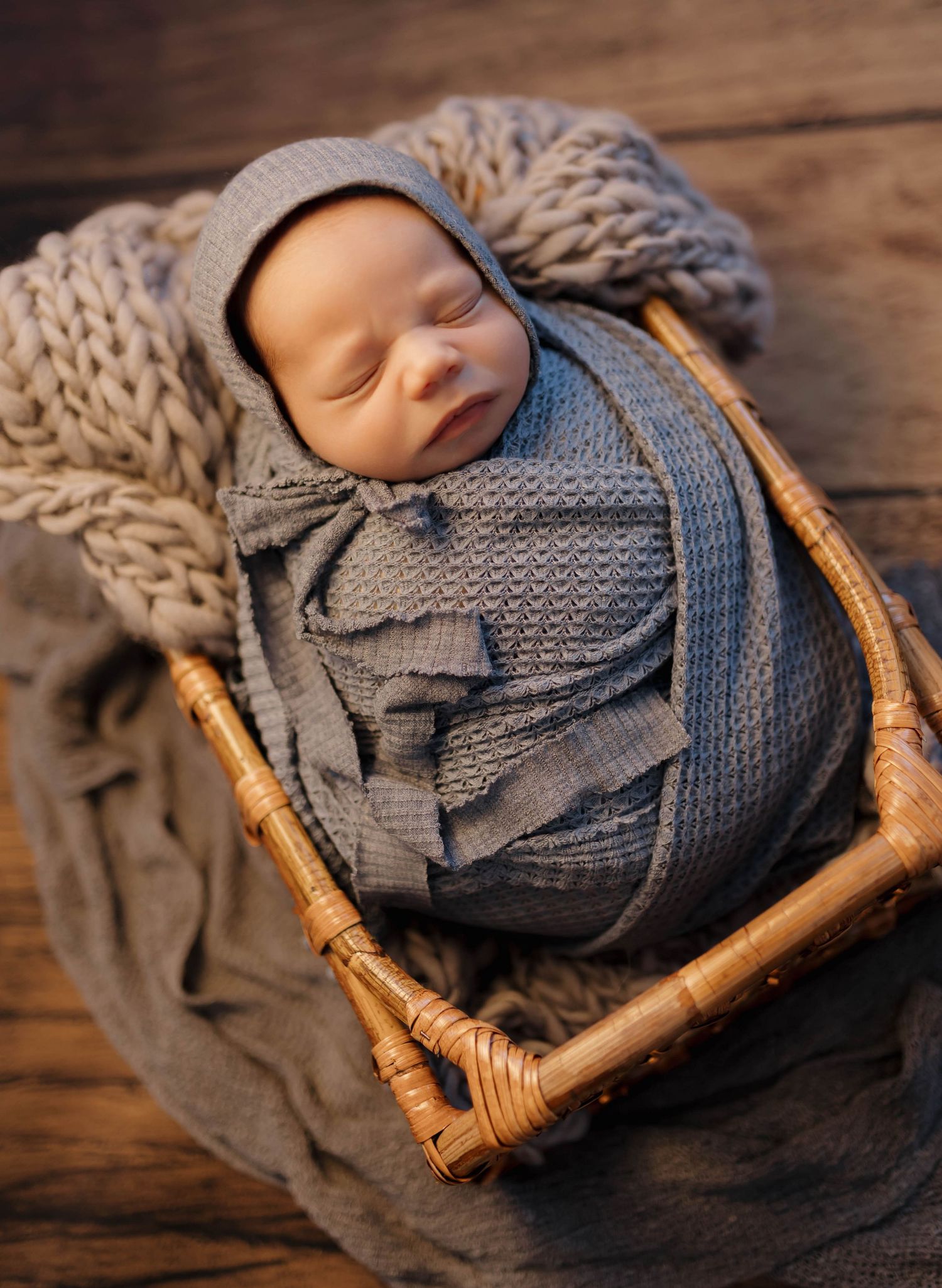 Sleeping newborn wrapped in cozy gray knit blanket rests in vintage wicker basket on rustic wooden surface.