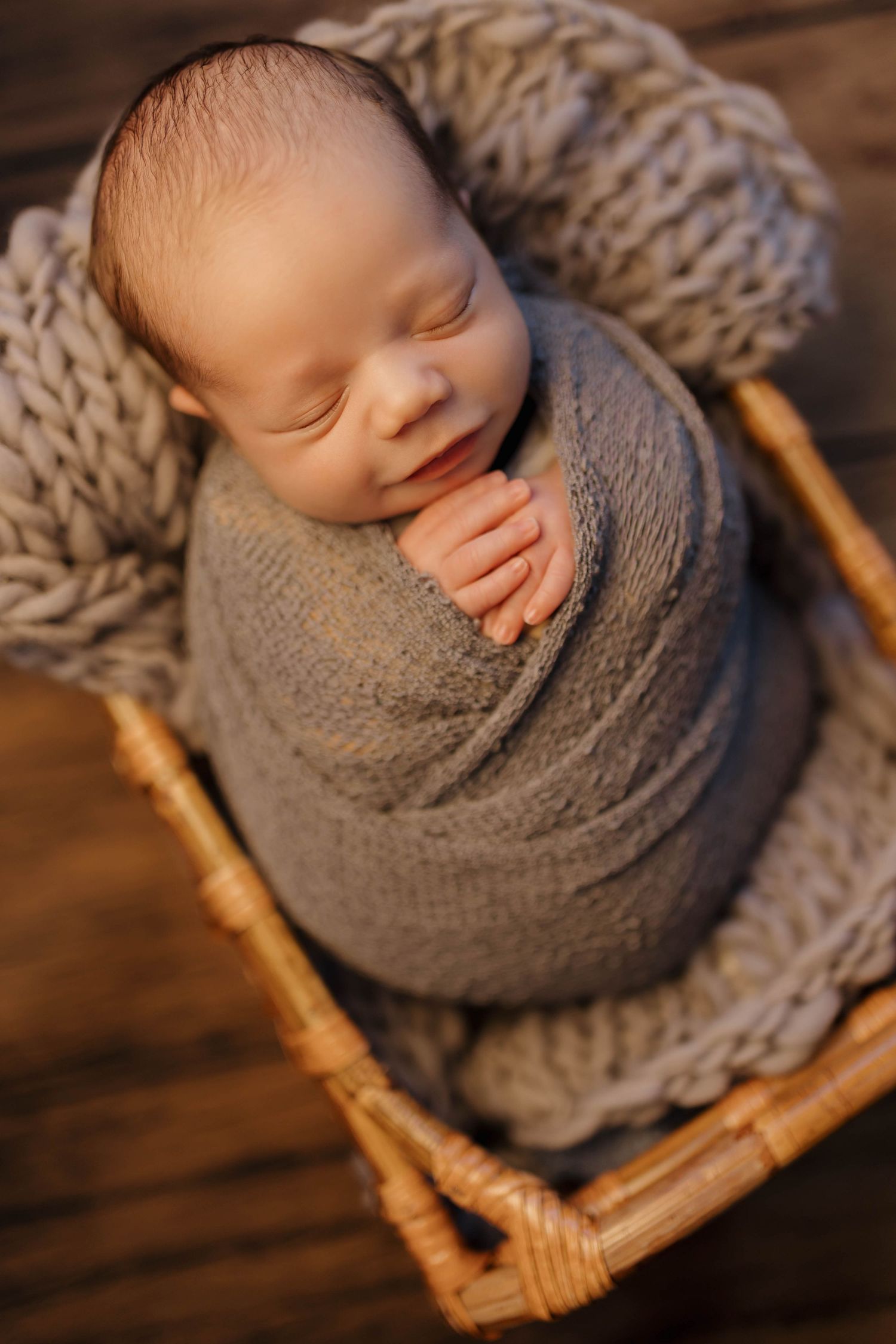 Peaceful newborn swaddled in soft gray knit wrap sleeps soundly in woven basket on wooden backdrop.