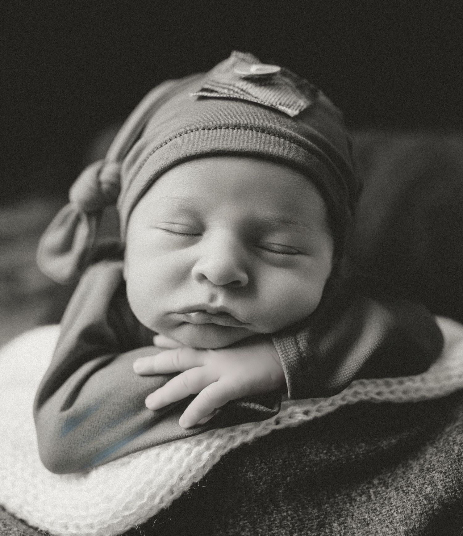 Peaceful sleeping newborn wearing a soft knit hat rests hands under chin in black and white portrait.