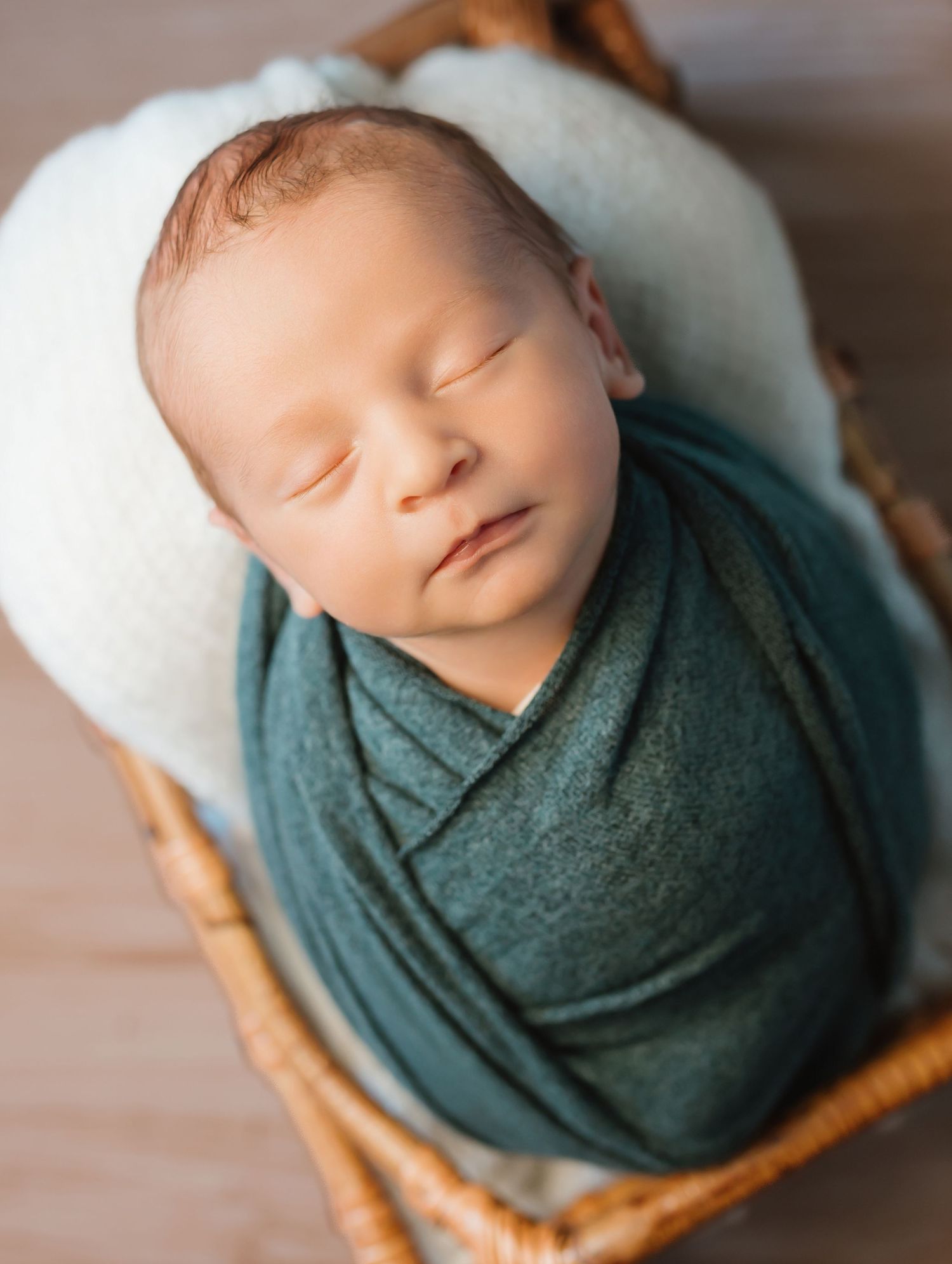 A sleeping newborn wrapped in a teal swaddle blanket rests peacefully in a wooden basket.