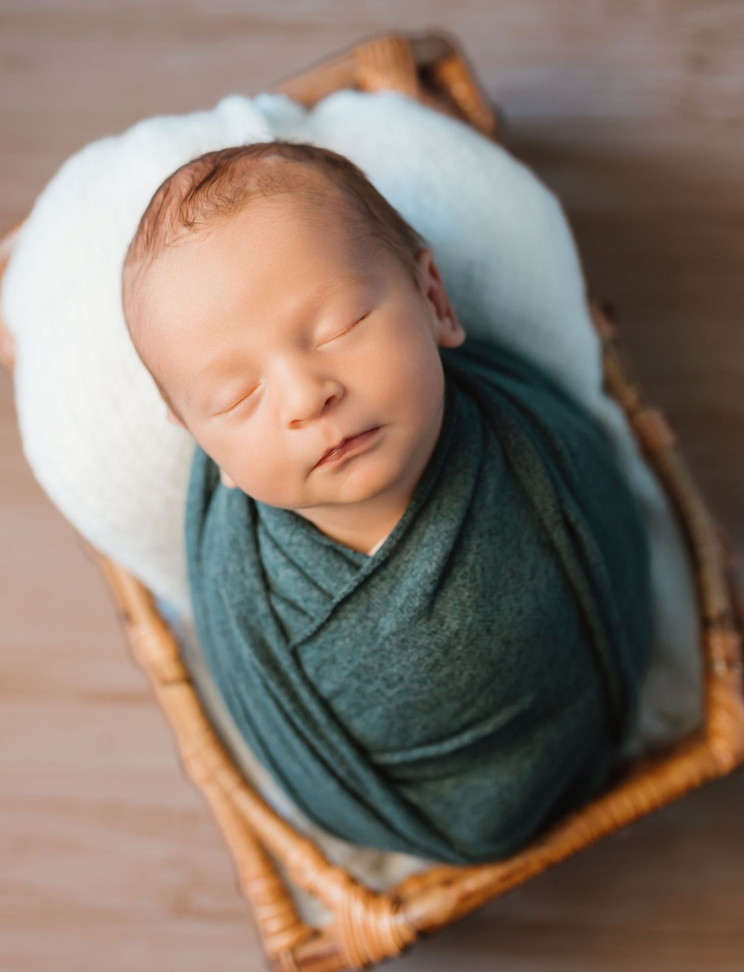 Newborn baby wrapped in teal swaddle cloth resting peacefully in wooden basket during professional photoshoot.