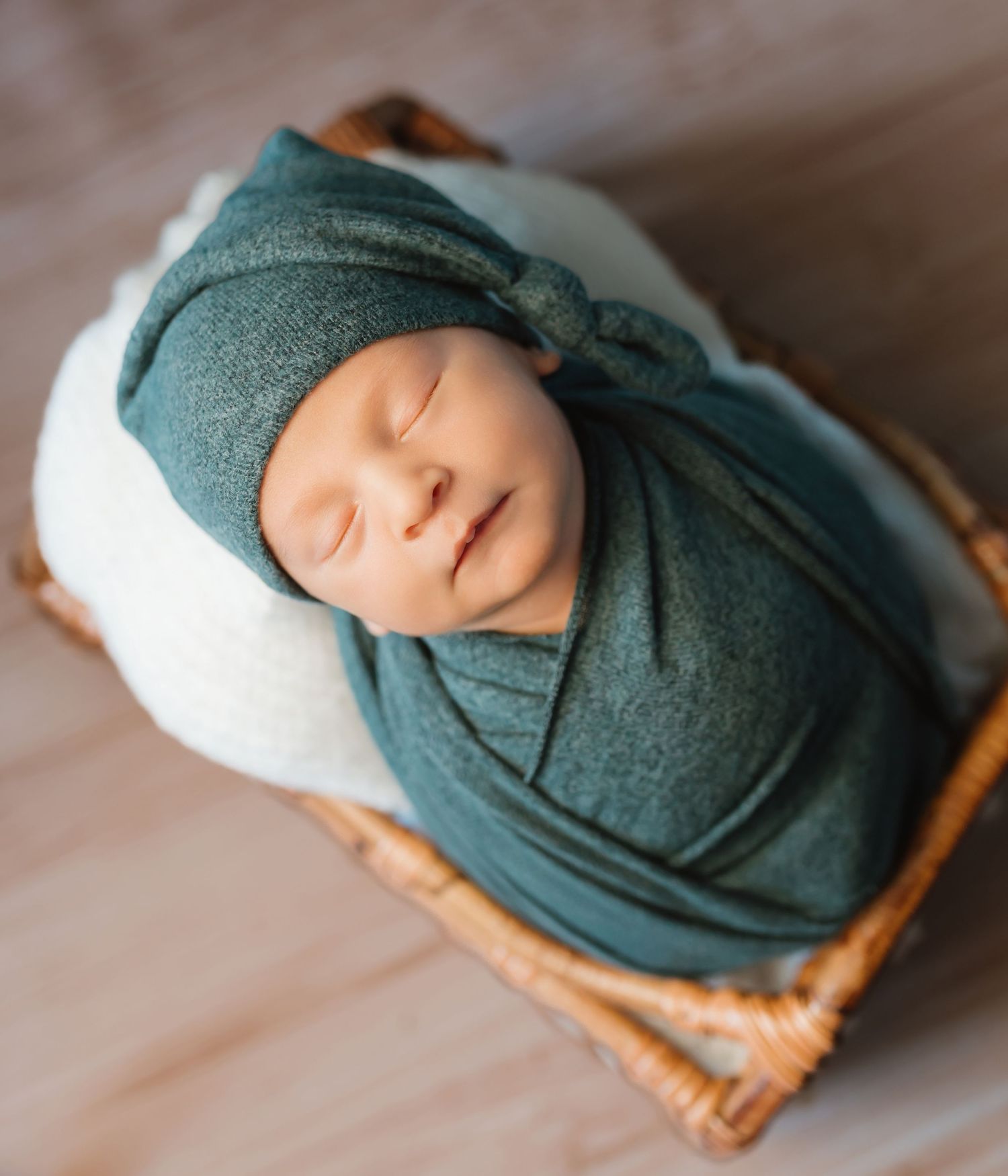 A newborn baby sleeping peacefully wrapped in a teal swaddle blanket on a cream-colored cushion.