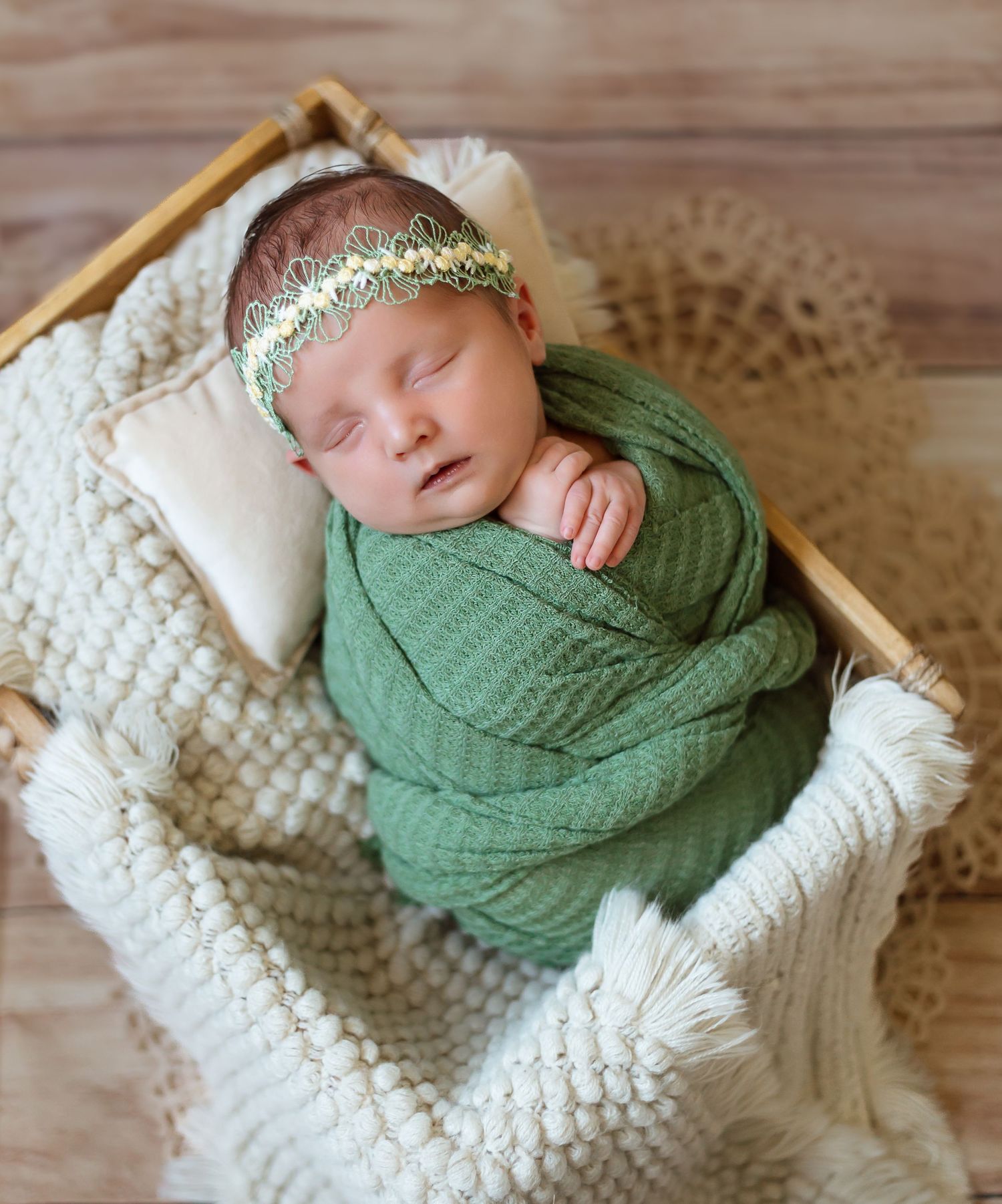 Sleeping newborn wrapped in green swaddle blanket resting on white textured knit blanket in wooden basket.
