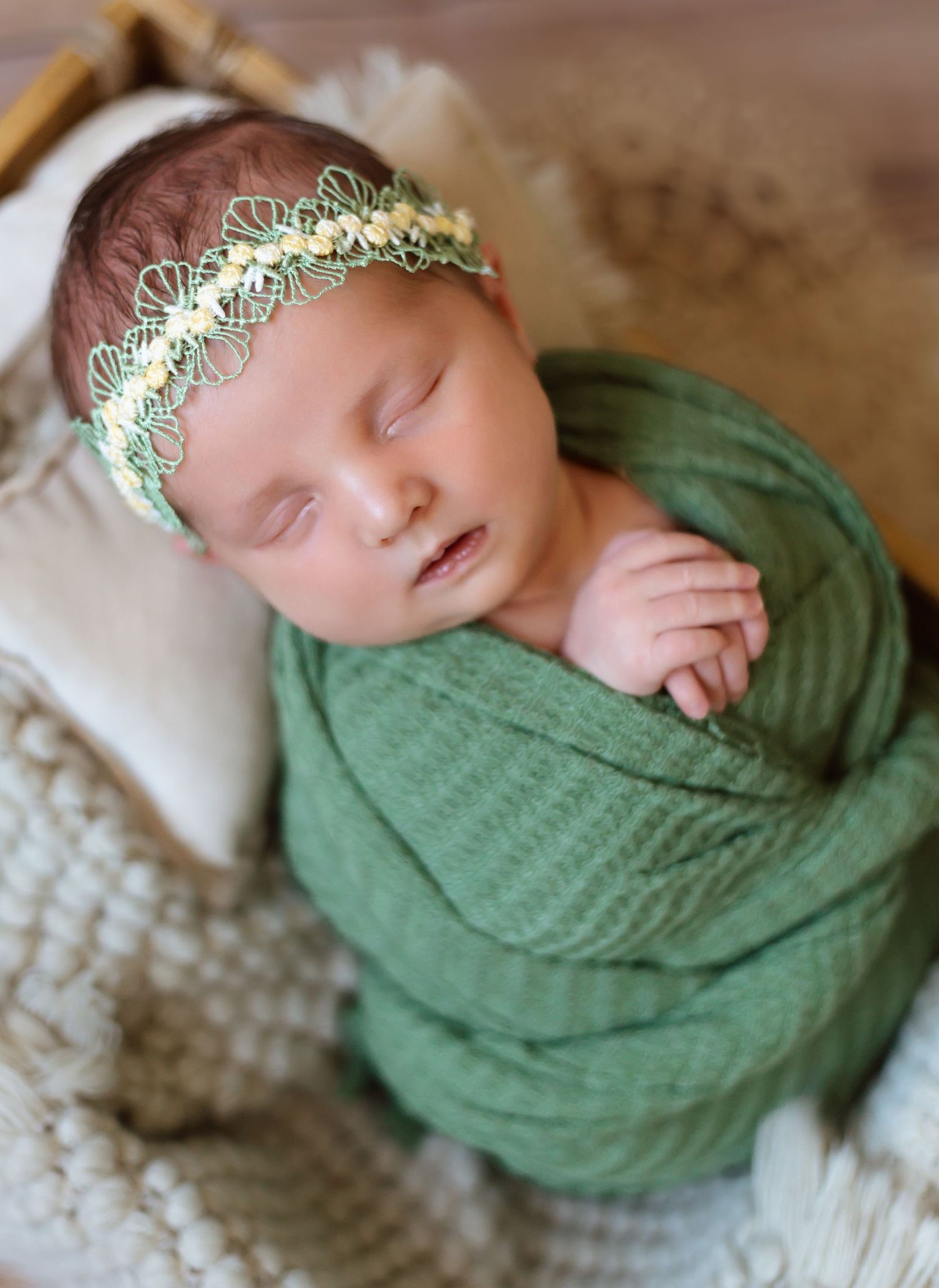 Newborn wrapped in soft green knit blanket with delicate lace headpiece resting on textured white backdrop.