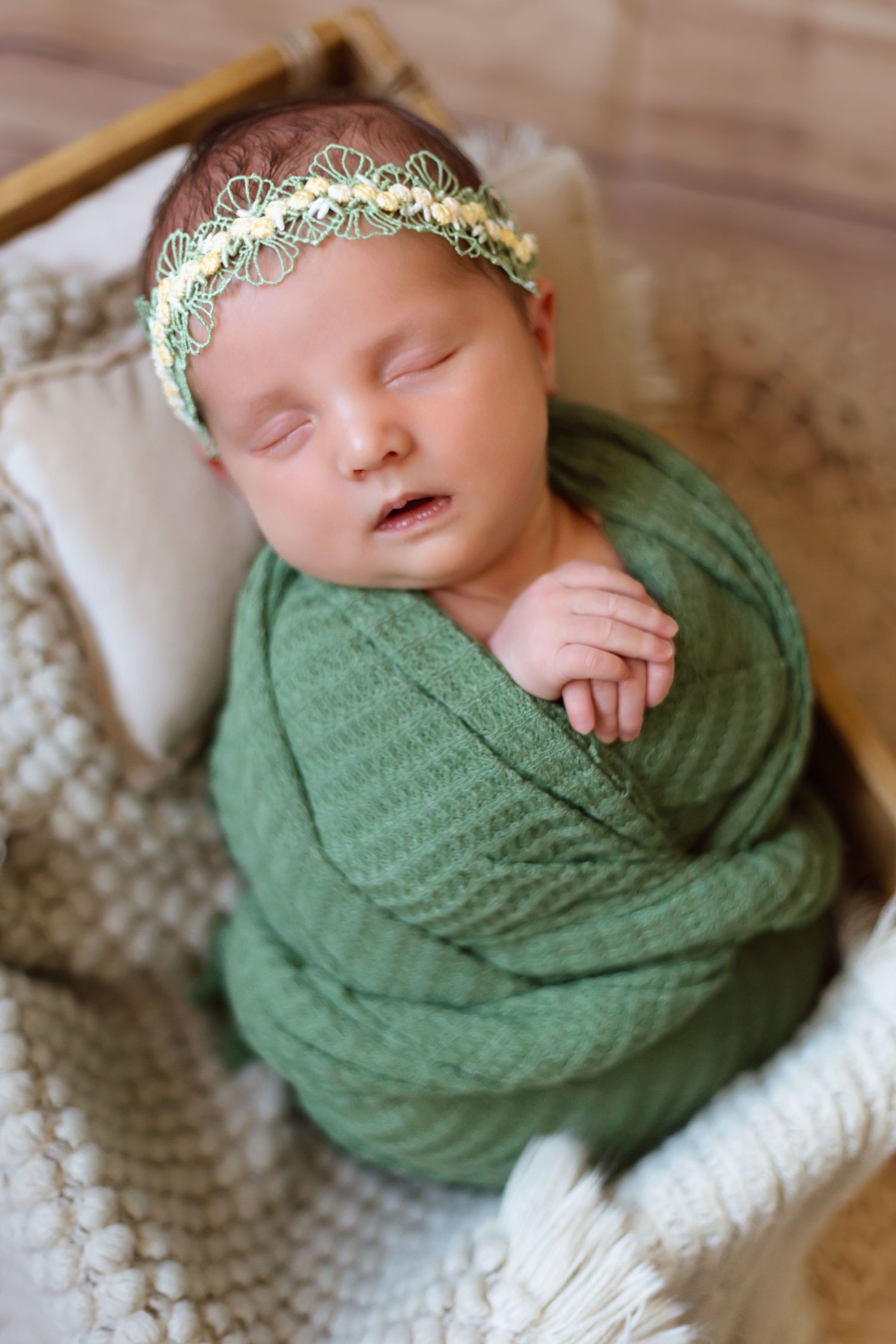 Sleeping newborn wrapped in soft green swaddle with delicate crown headpiece resting on textured blanket.