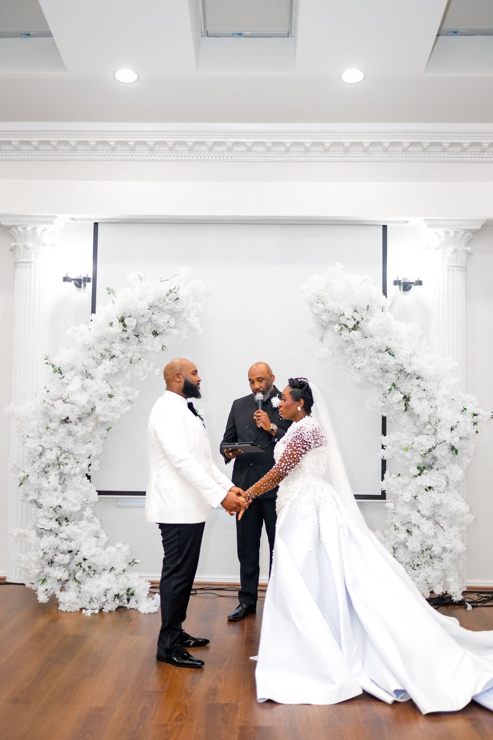 A wedding ceremony unfolds in a bright white room adorned with elegant white floral arrangements.