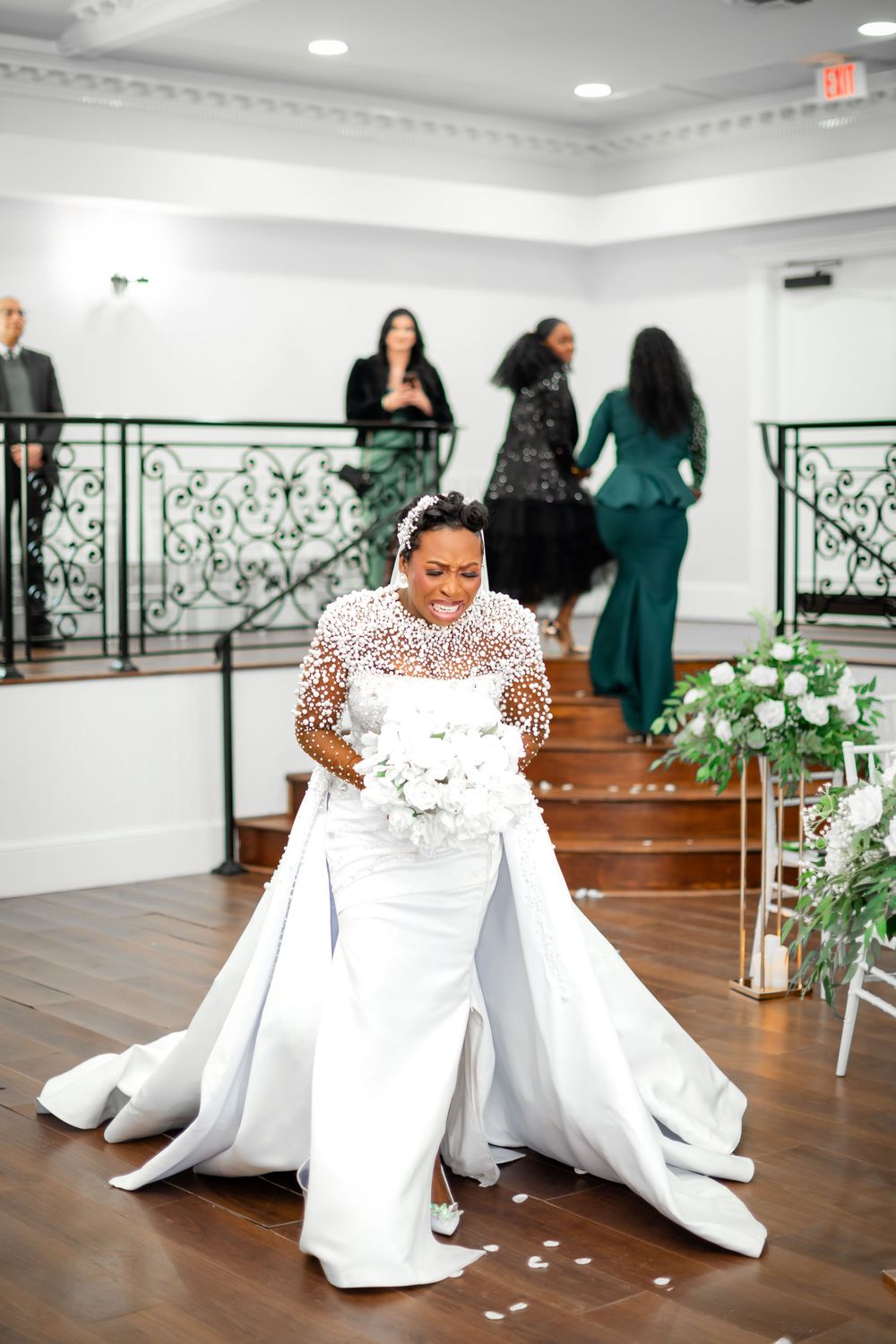 A person in a white wedding dress sits on the floor during a joyful moment at an indoor venue with hardwood floors.