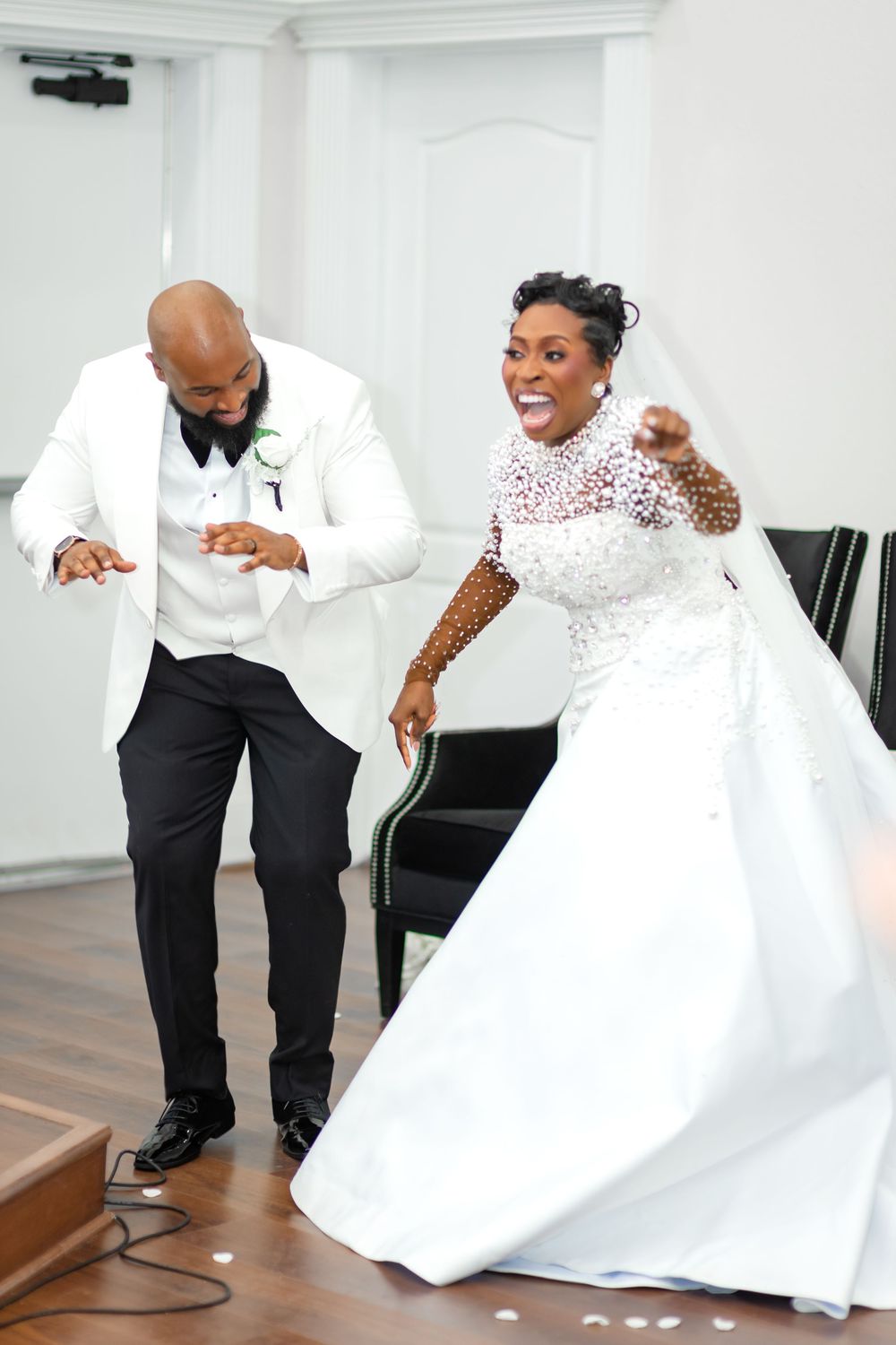 A couple shares a playful dance moment on hardwood floors in a white-walled venue with elegant decor.