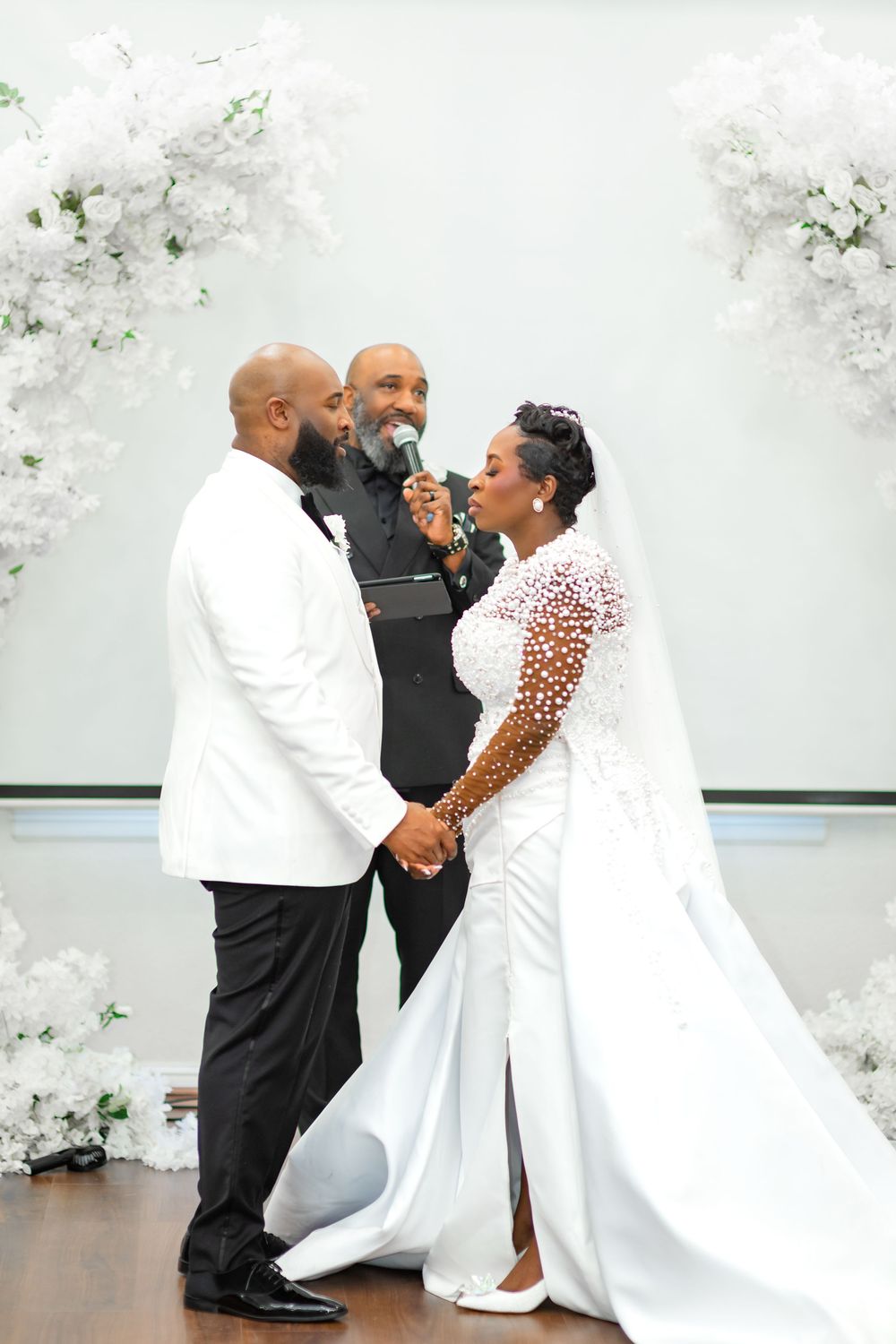 A joyful wedding ceremony with a couple in elegant white attire sharing a moment with their officiant against floral decor.