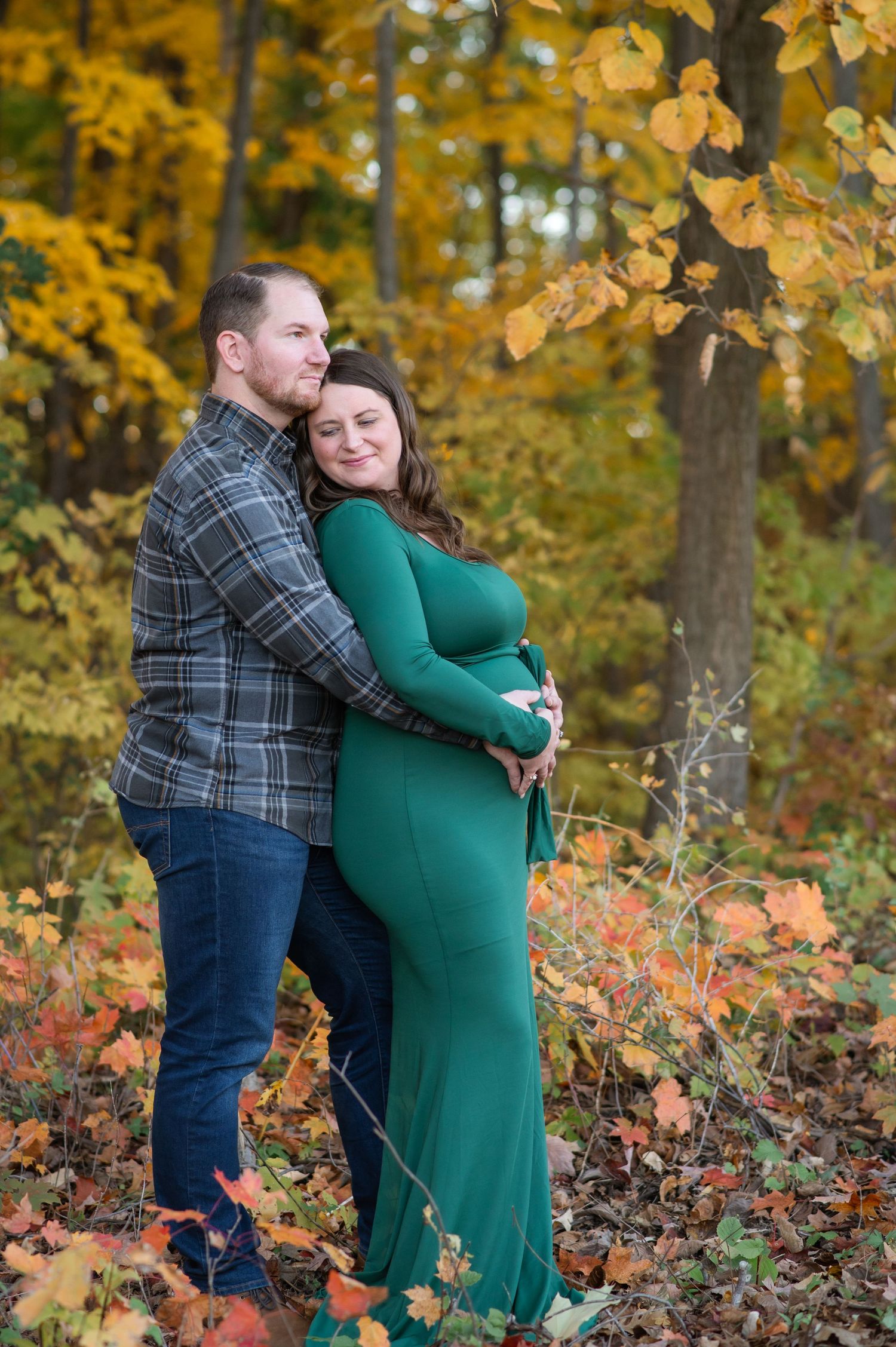 A couple embraces during an autumn maternity photo shoot surrounded by golden fall foliage in the woods.