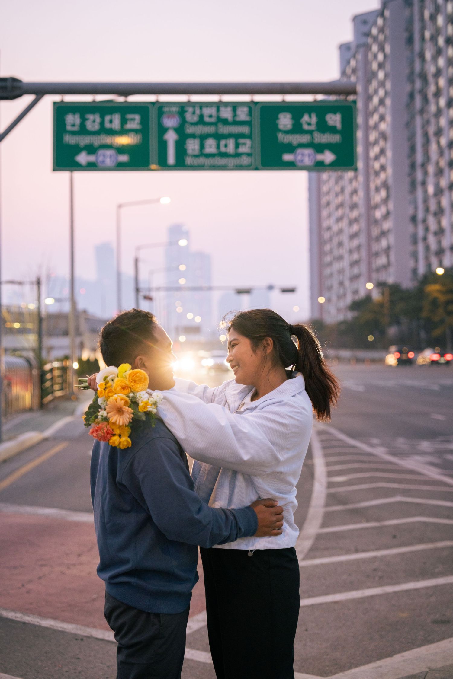 Two figures embrace on an empty urban street at dusk with Korean street signs visible in the background.