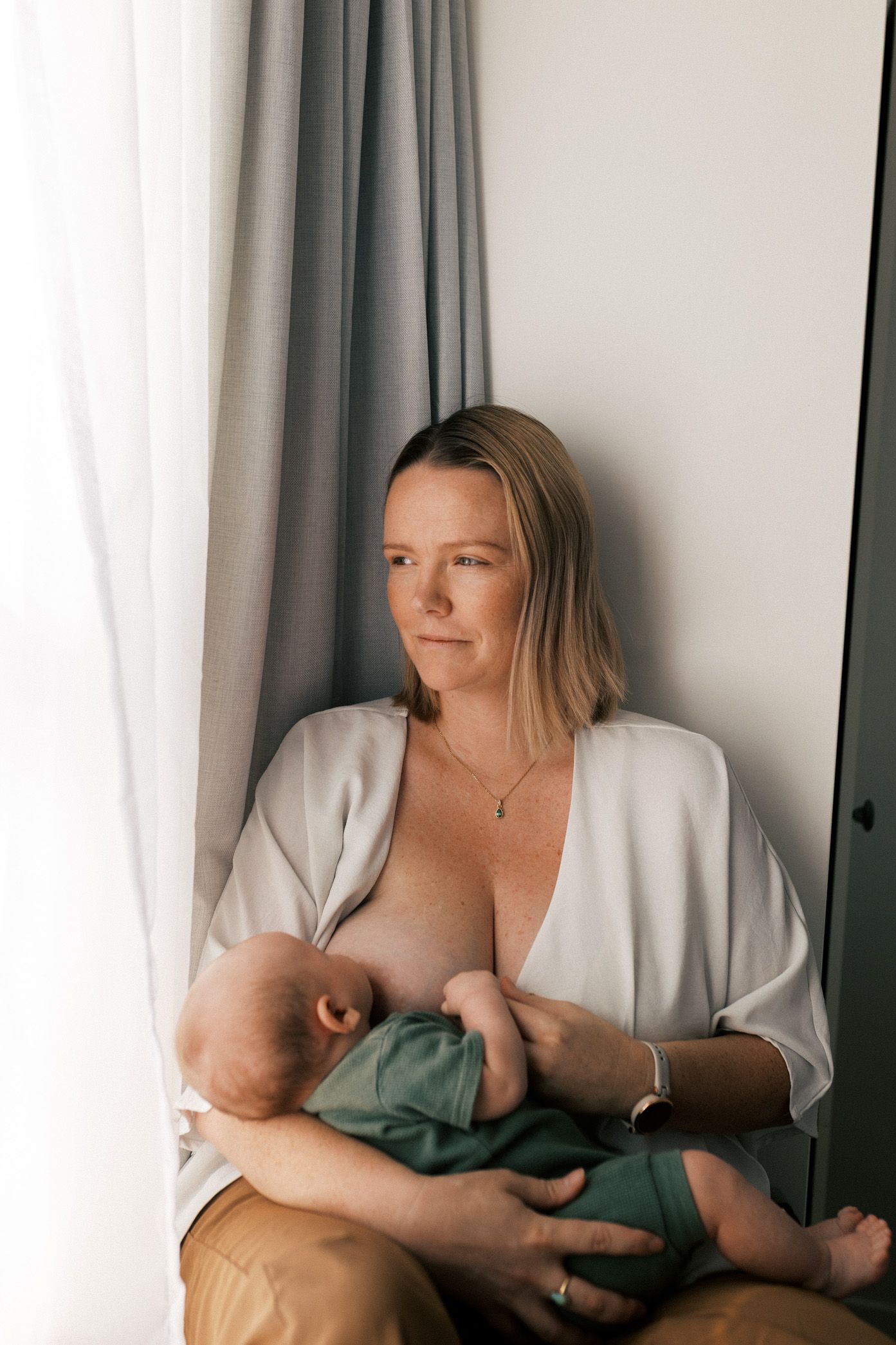 A tender moment captured in soft natural light as a parent nurses their baby near a window with white curtains.