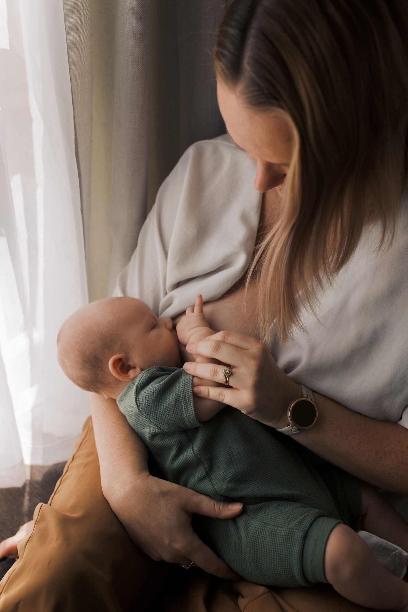 A tender moment of breastfeeding as a baby in green clothing nurses while being cradled near a window.