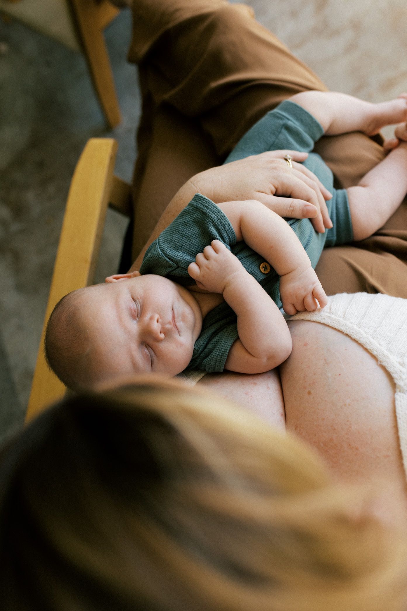 Sleeping baby in teal outfit resting peacefully on parent's lap in warm natural lighting.