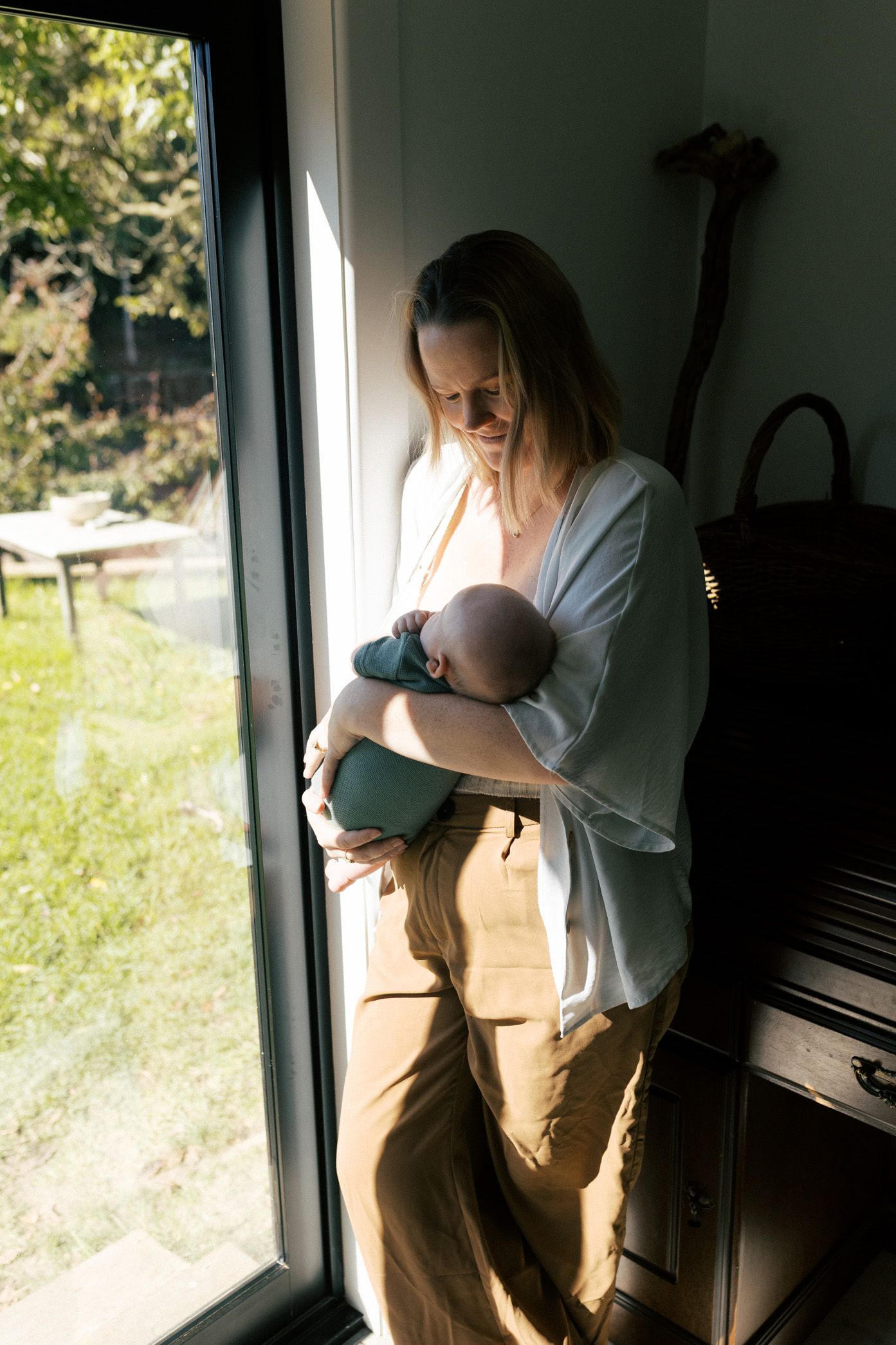 A mother in a light shirt and brown pants stands by a window while nursing her baby in a series of connected photos.