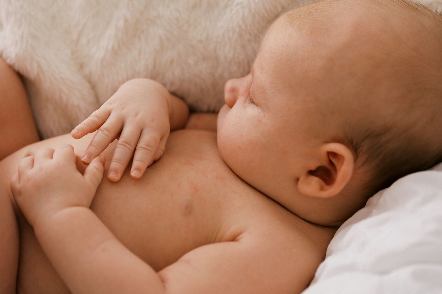 A baby lies peacefully sleeping on soft white bedding while a gentle hand caresses their back.