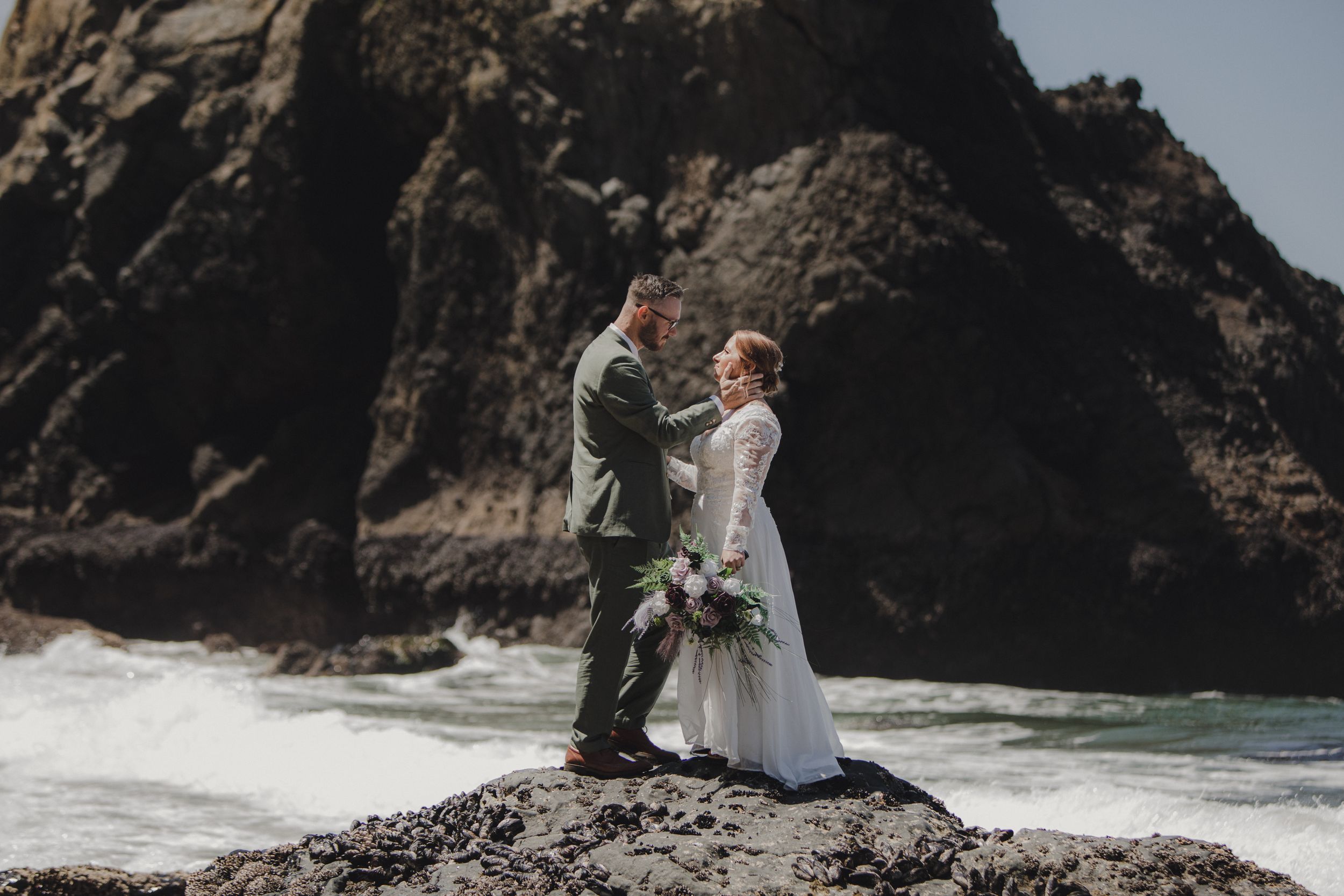 Couple in wedding attire embracing on dramatic rocky beach coastline with crashing waves and cliffs in background.