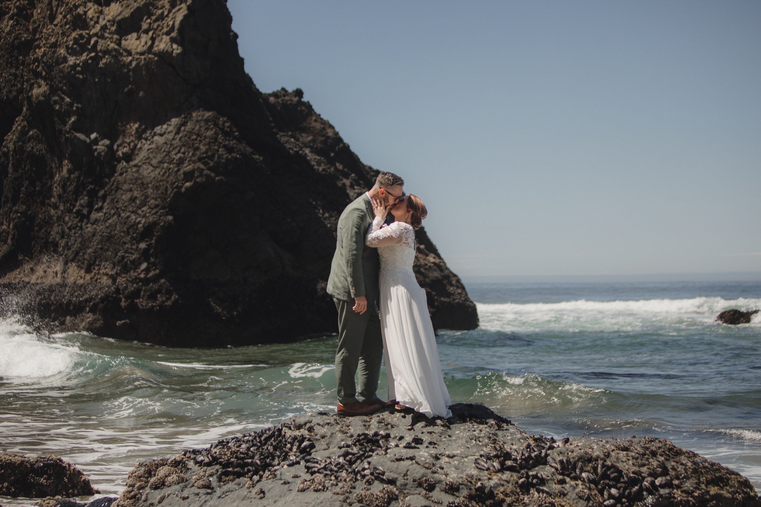 A couple in wedding attire embraces on rocky coastal cliffs with crashing waves and ocean views in the background.
